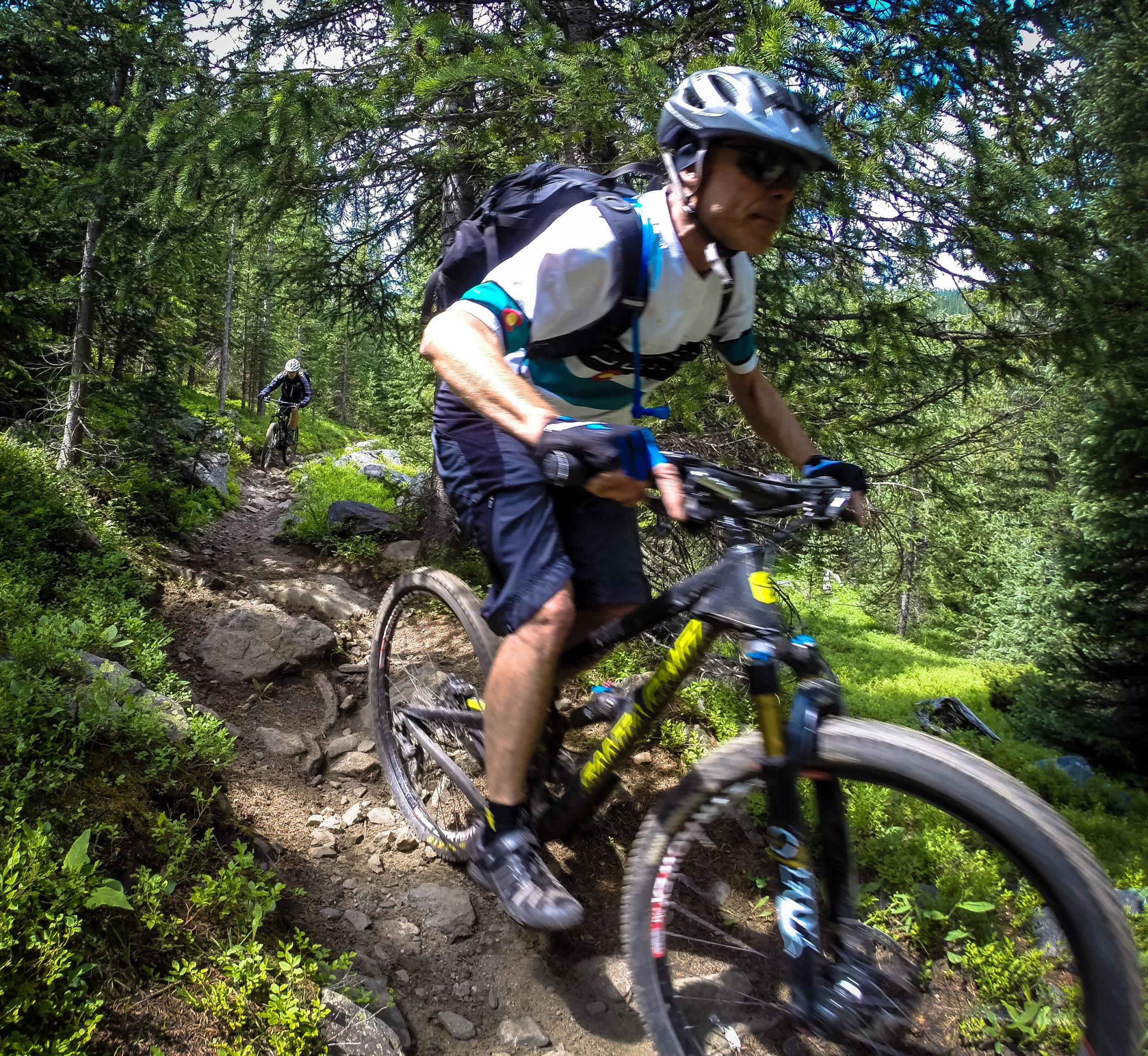 A mountain biker navigating a rocky trail in a forested area, with another cyclist visible in the background. The scene captures the dynamic action of mountain biking on a sunny day, surrounded by trees and greenery. Green's Creek mountain bike trail.