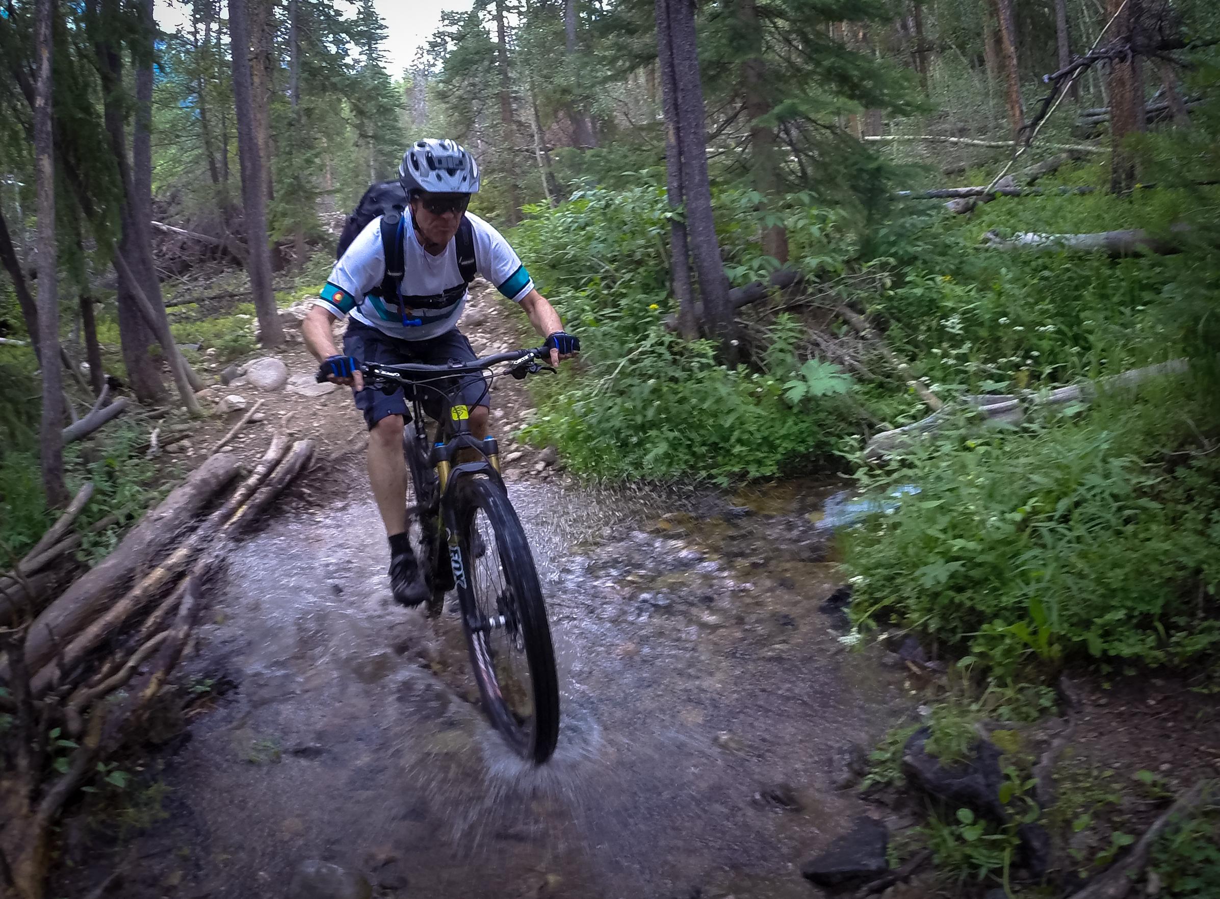 A person riding a mountain bike through a shallow stream in a forested area, surrounded by trees and greenery. The rider is wearing a helmet and casual outdoor clothing, slightly splashing water as they navigate the trail. Green's Creek mountain bike trail.