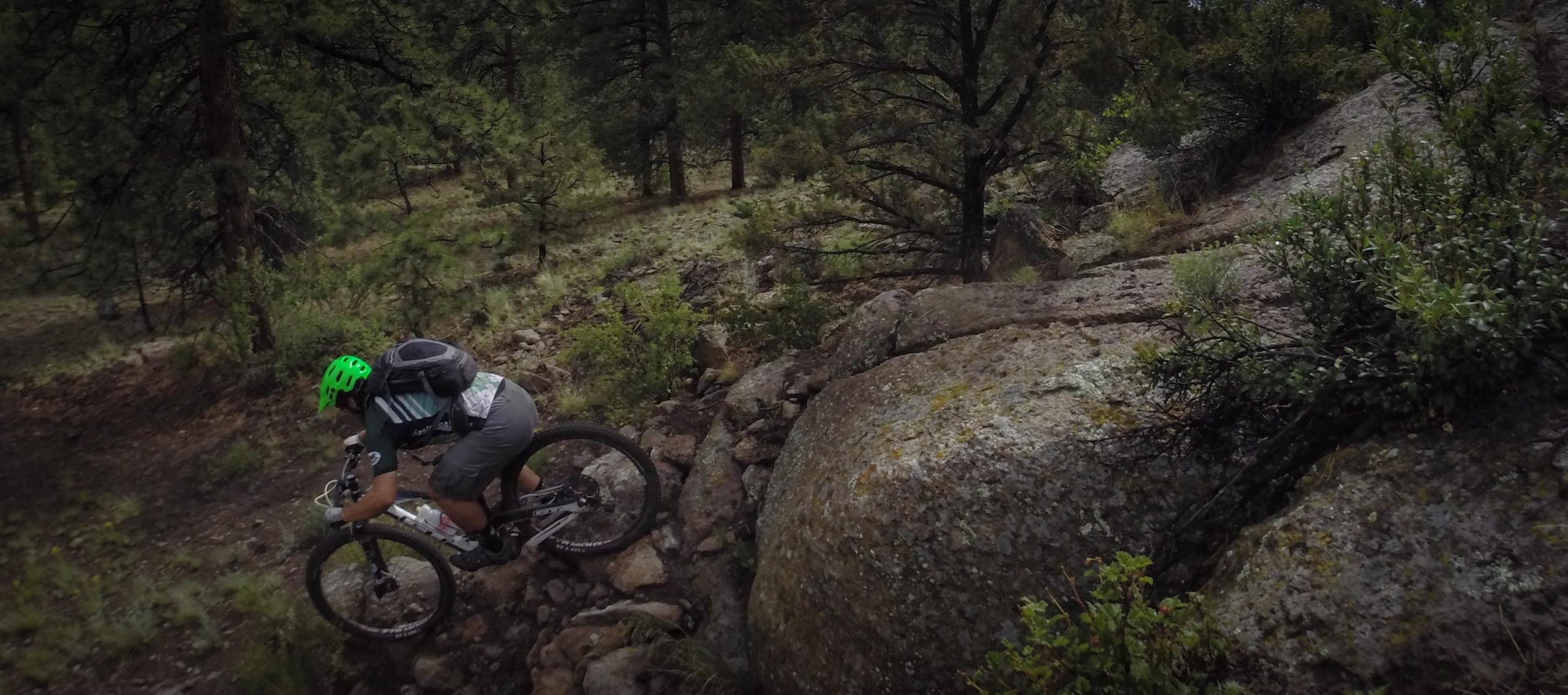 A mountain biker navigates a rocky trail surrounded by dense trees and vegetation. The rider, wearing a green helmet and a backpack, leans into the bike as they tackle the rough terrain. Large boulders and scattered rocks are visible along the path, highlighting the challenging nature of the ride. Cottonwood mountain bike trail.