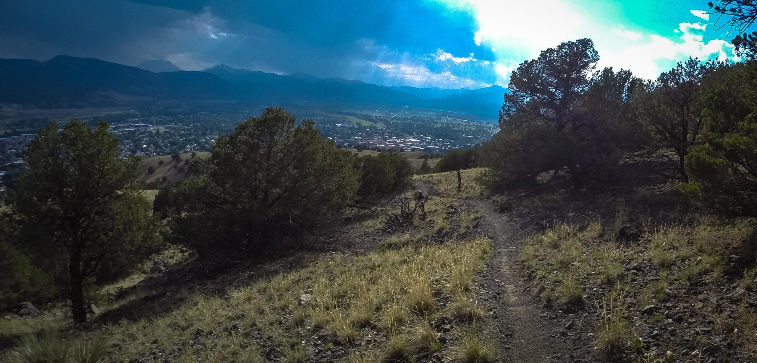 A scenic view from a hillside trail, featuring a winding dirt path surrounded by low vegetation and trees. In the background, a valley unfolds with a small town and mountains visible under a dramatic sky with varying shades of blue and gray. Cottonwood mountain bike trail.