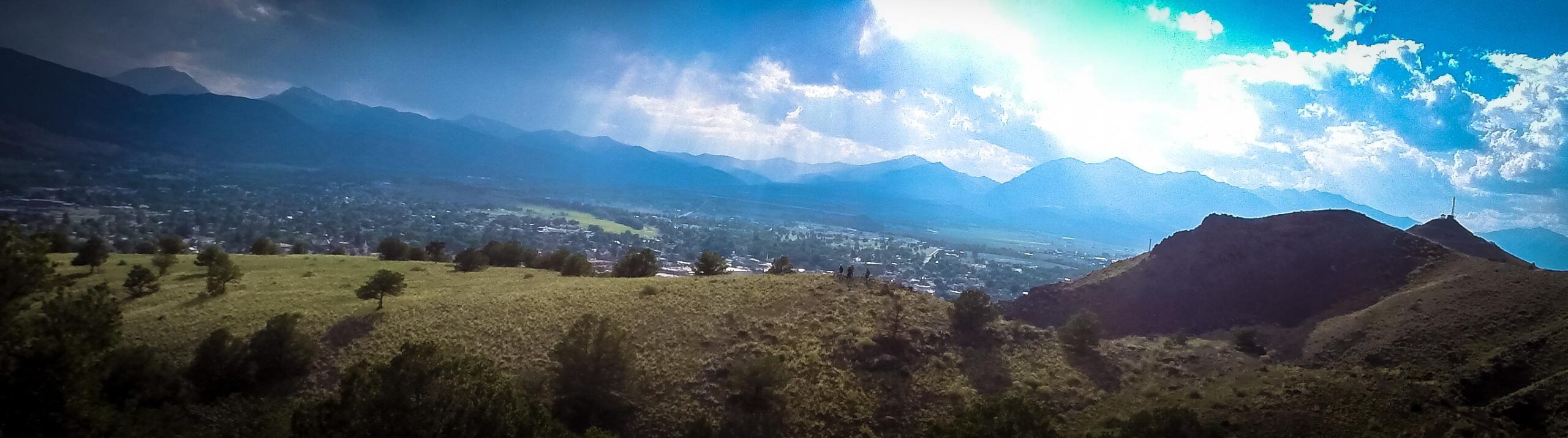 A panoramic view of rolling green hills leading to distant mountains, bathed in natural sunlight under a partly cloudy sky. The landscape showcases a vibrant blend of grasslands and trees, with a small town visible in the valley below, creating a serene outdoor scene. Arkansas Hills mountain bike trail.