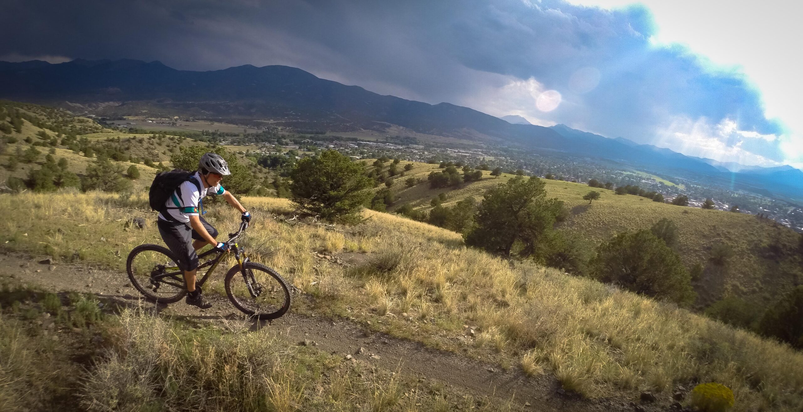 A mountain biker navigates a rugged trail on a hillside, surrounded by rolling green terrain and distant mountains under a partly cloudy sky. The scene captures a sense of adventure and outdoor activity in nature. Arkansas Hills mountain bike trail.