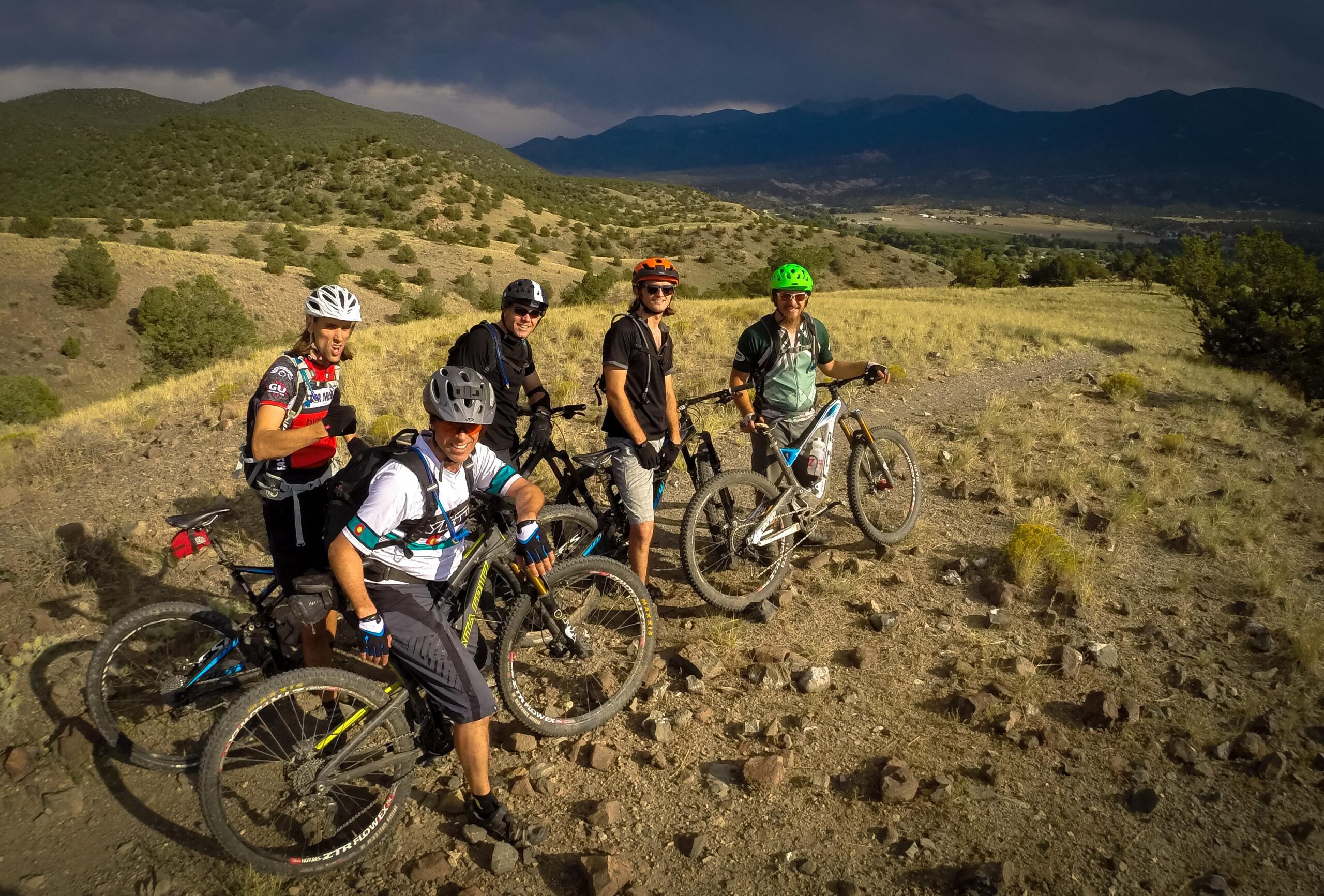 A group of five mountain bikers standing on a rocky trail, surrounded by hilly terrain and sparse vegetation. They are wearing cycling gear and helmets, with their bikes positioned beside them. The background features mountains under a partly cloudy sky. Arkansas Hills mountain bike trail.