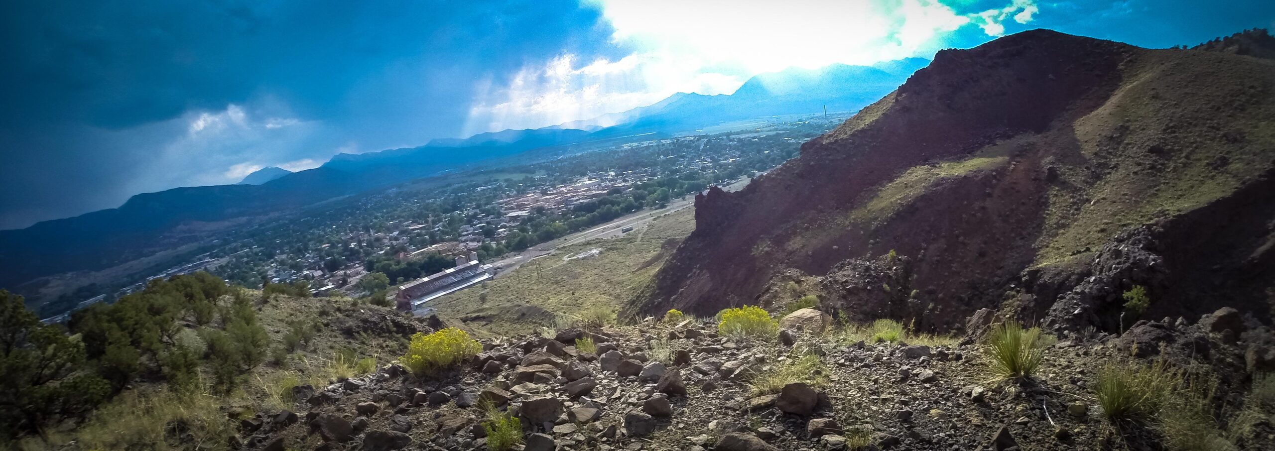 A panoramic view of a mountainous landscape with dramatic clouds overhead. In the foreground, rocky terrain and sparse vegetation are visible, leading down to a small town nestled in the valley. The distant mountains rise under the cloudy sky, creating a stunning backdrop for the scene. Arkansas Hills mountain bike trail.
