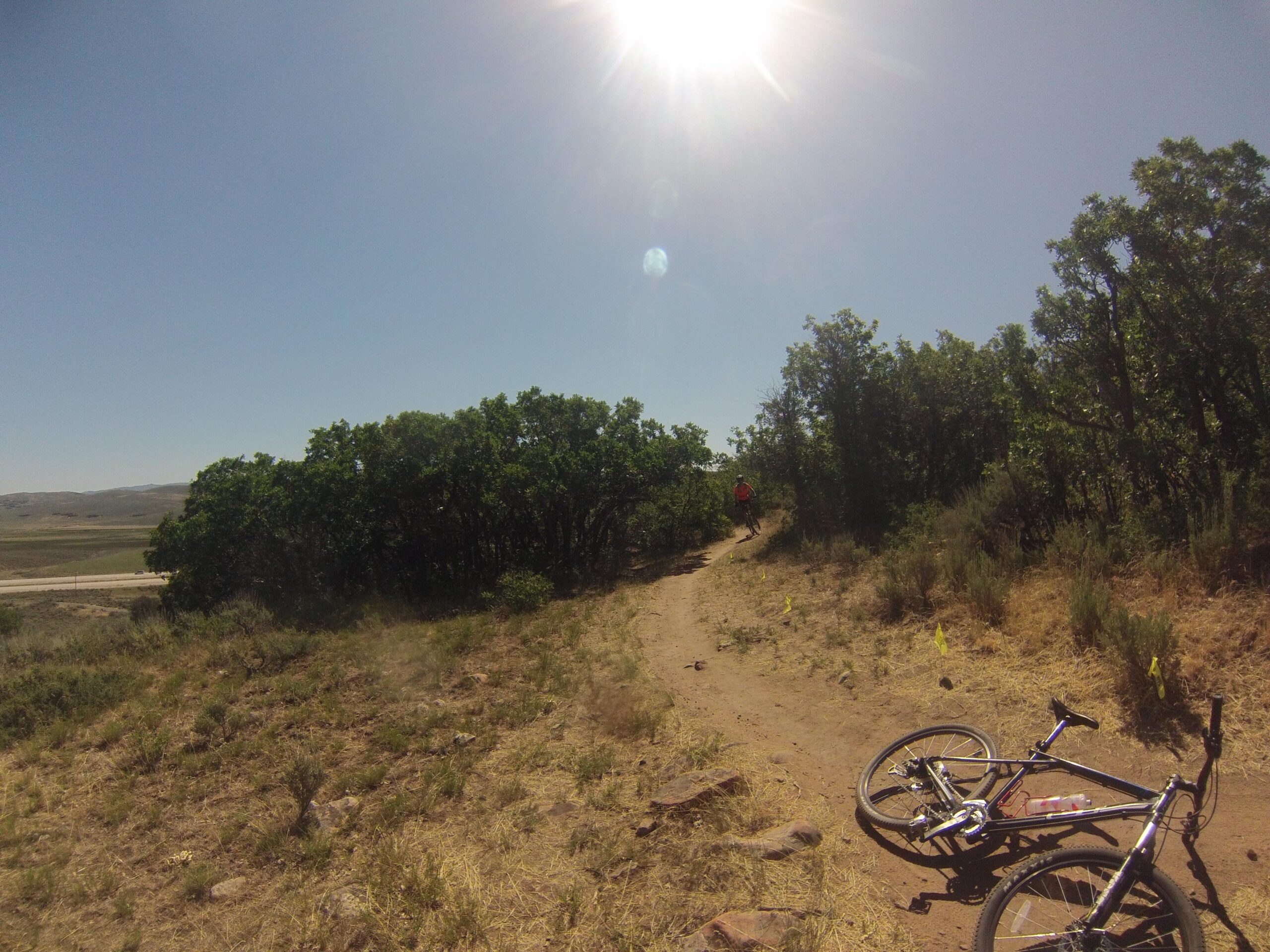 A sunlit trail winding through greenery, with a mountain bike lying on the ground in the foreground. A cyclist in a red shirt can be seen riding along the path in the background. Clear blue sky and open landscape stretch out in the distance. Round Valley mountain bike trail.