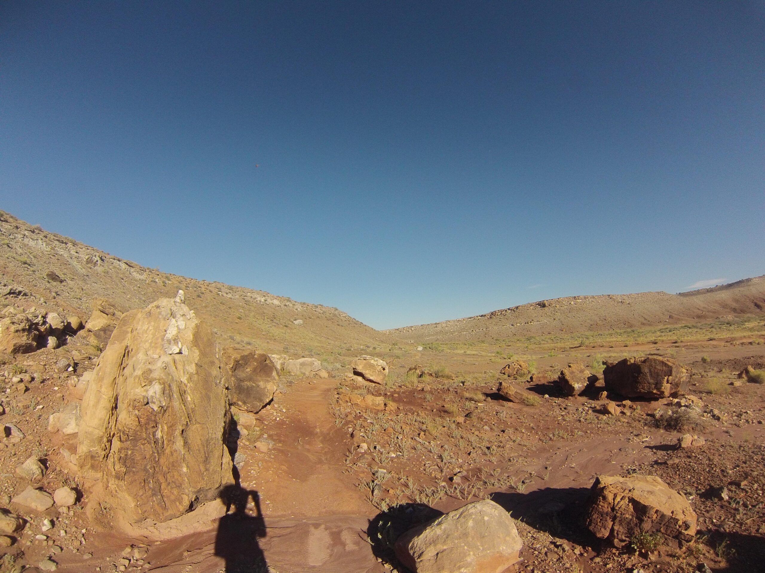Alt text: A rocky landscape under a clear blue sky, featuring large boulders and a dirt path winding through a dry, rugged terrain. Shadow of a person taking a photo in the foreground. Klondike Bluffs mountain bike trail.