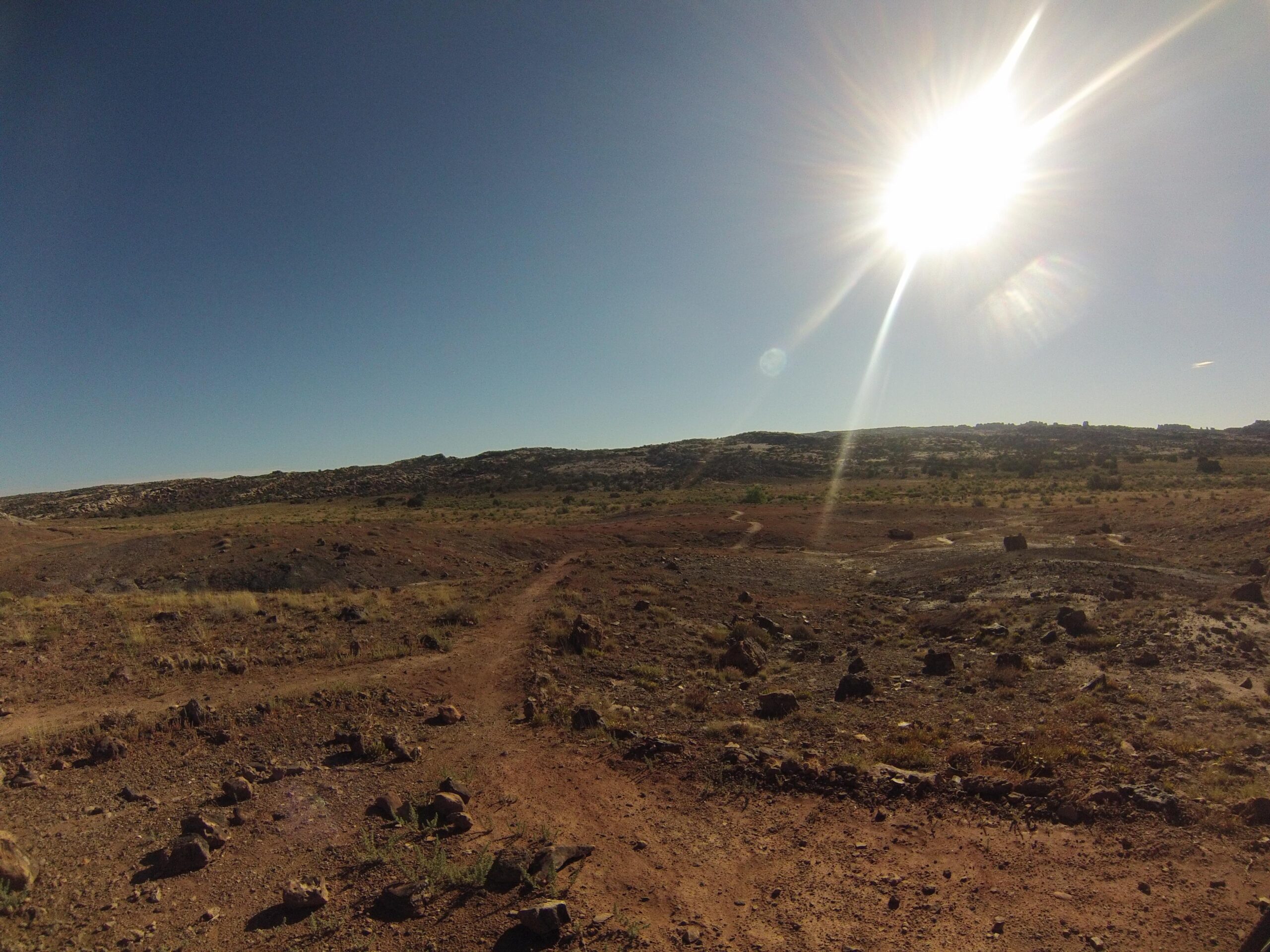 A sunlit landscape featuring a clear blue sky above a dry, rocky terrain. The foreground shows a dirt path winding through sparse vegetation and scattered rocks, leading towards a distant hillside. Klondike Bluffs mountain bike trail.