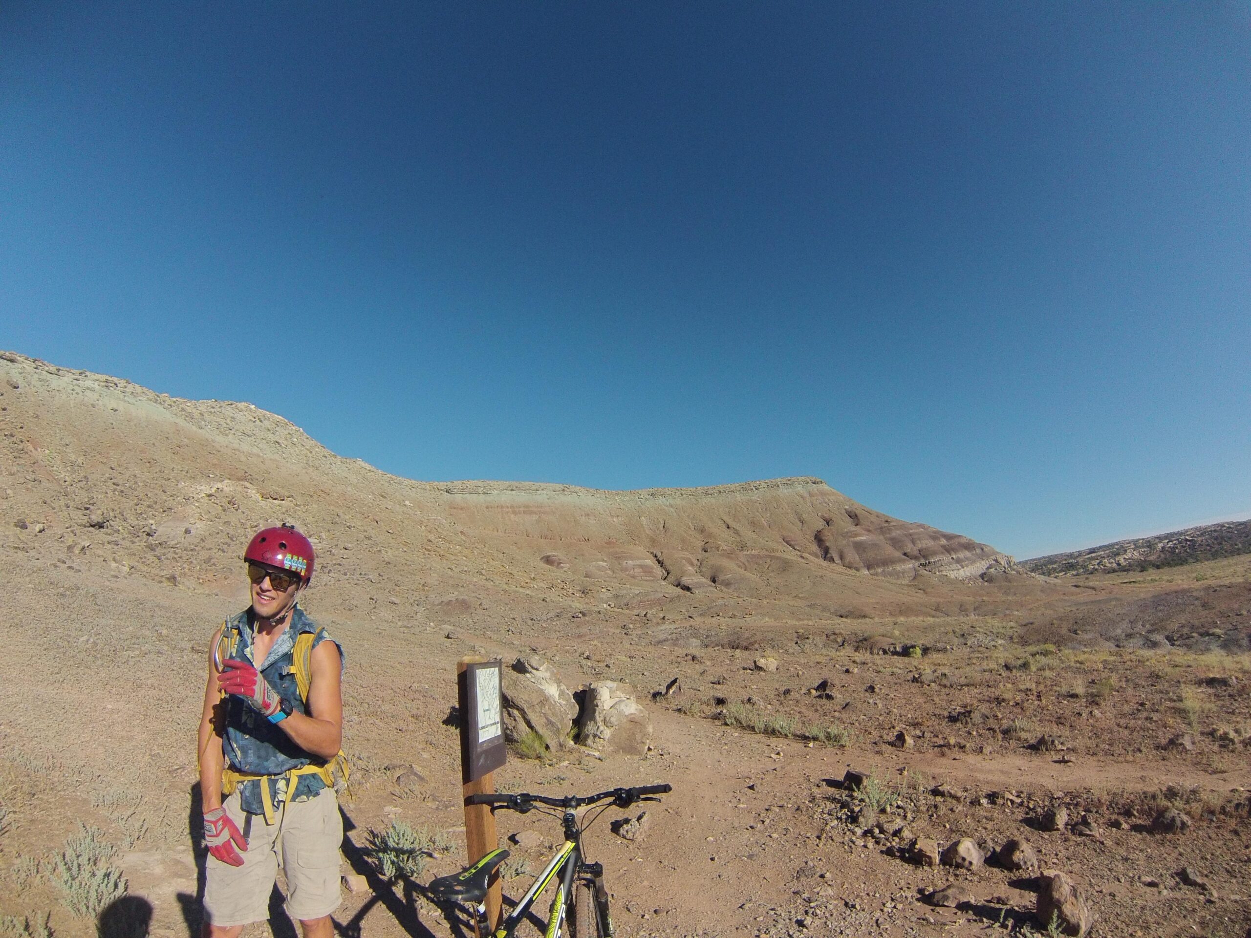 A person wearing a red helmet and gloves stands beside a mountain bike at a trailhead in a rocky, dry landscape. In the background, the terrain features layered hills under a clear blue sky. Klondike Bluffs mountain bike trail.
