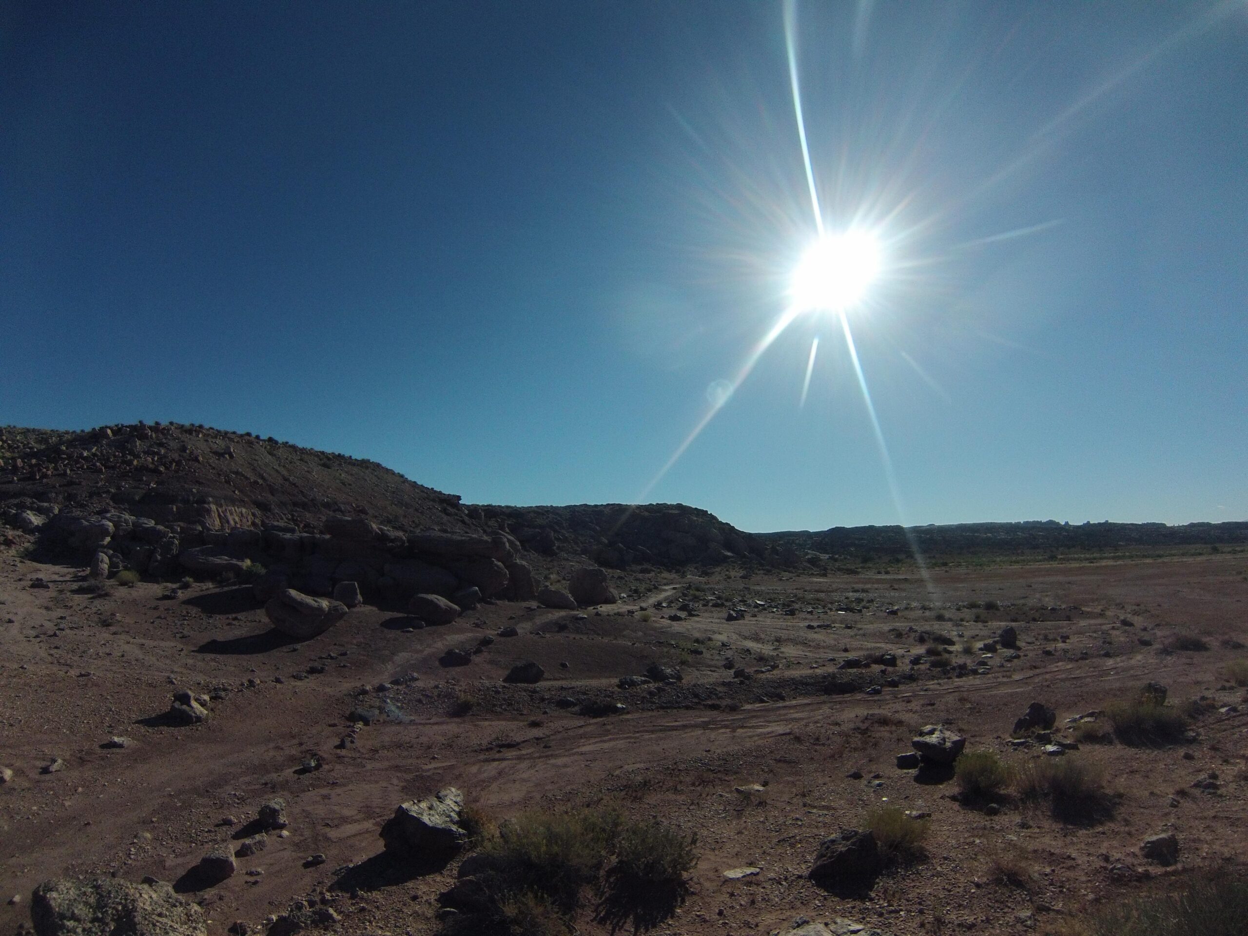 Desert landscape under a bright sun, featuring rugged terrain with scattered rocks and sparse vegetation. Clear blue sky above with a hint of distant hills. Klondike Bluffs mountain bike trail.