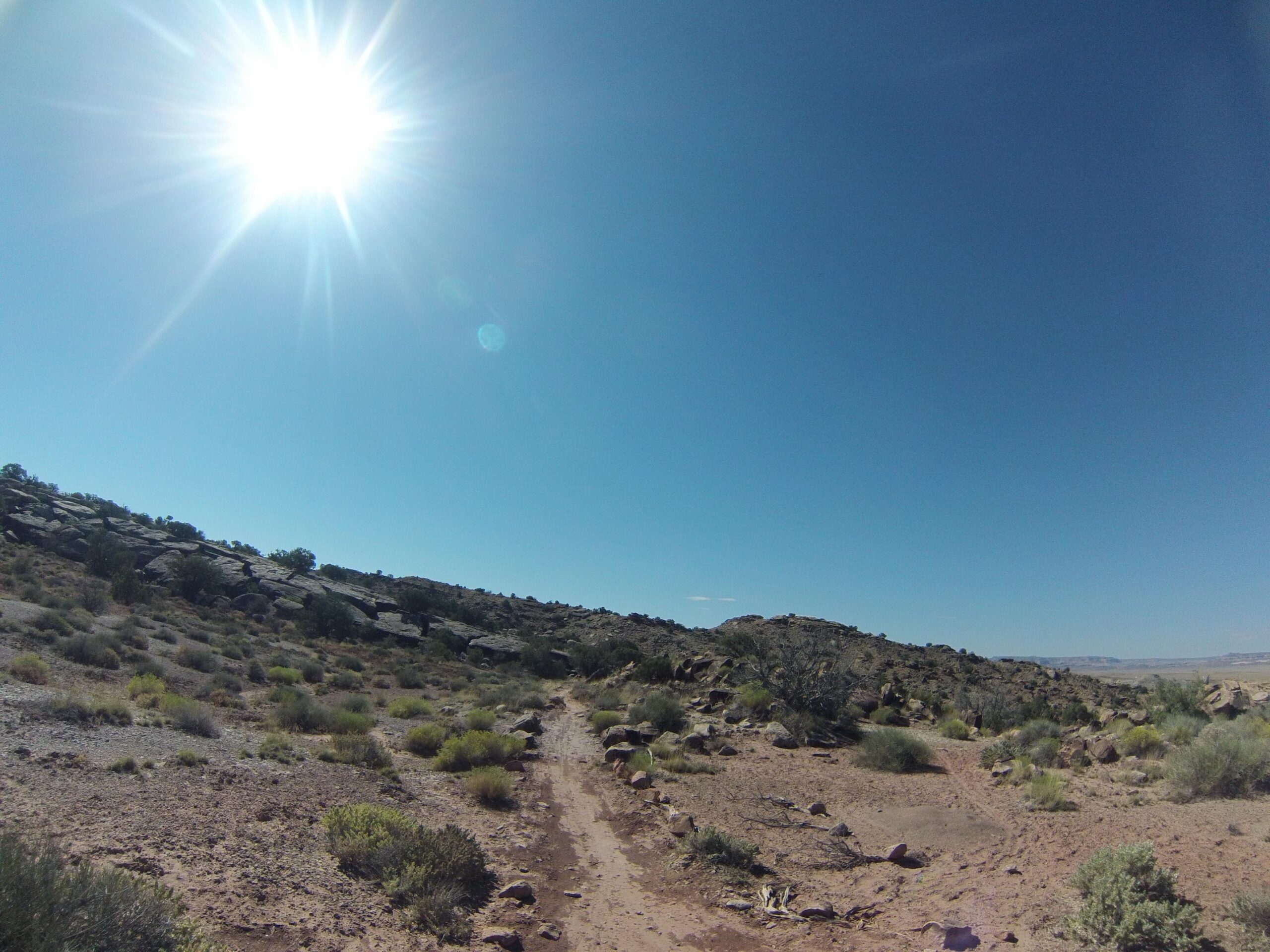 A sunlit landscape featuring a dirt path winding through a desert-like terrain, with sparse vegetation and rocky hills under a bright blue sky. The sun shines prominently in the upper left corner, illuminating the scene. Klondike Bluffs mountain bike trail.