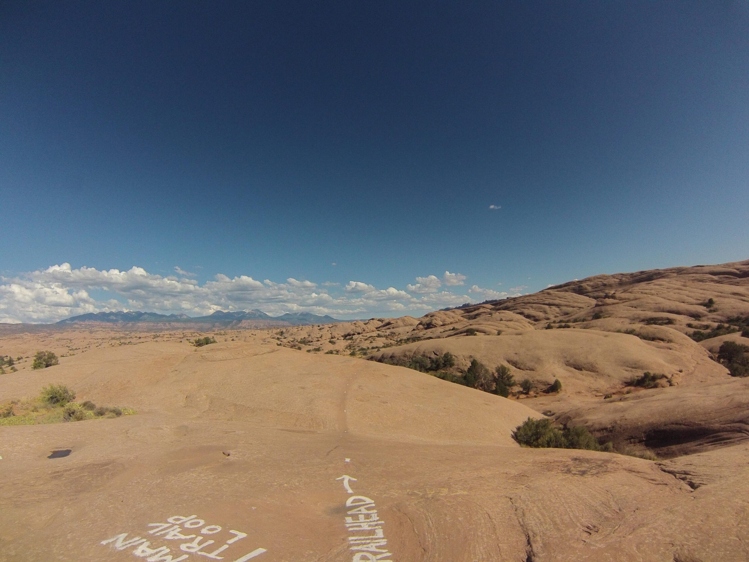 A panoramic view of a desert landscape featuring rolling sandstone hills under a bright blue sky with scattered clouds. A trail marker indicating "trailhead" is visible on the rocky surface in the foreground. The distant mountains are partially obscured by clouds, creating a picturesque and serene outdoor scene. Slickrock mountain bike trail.