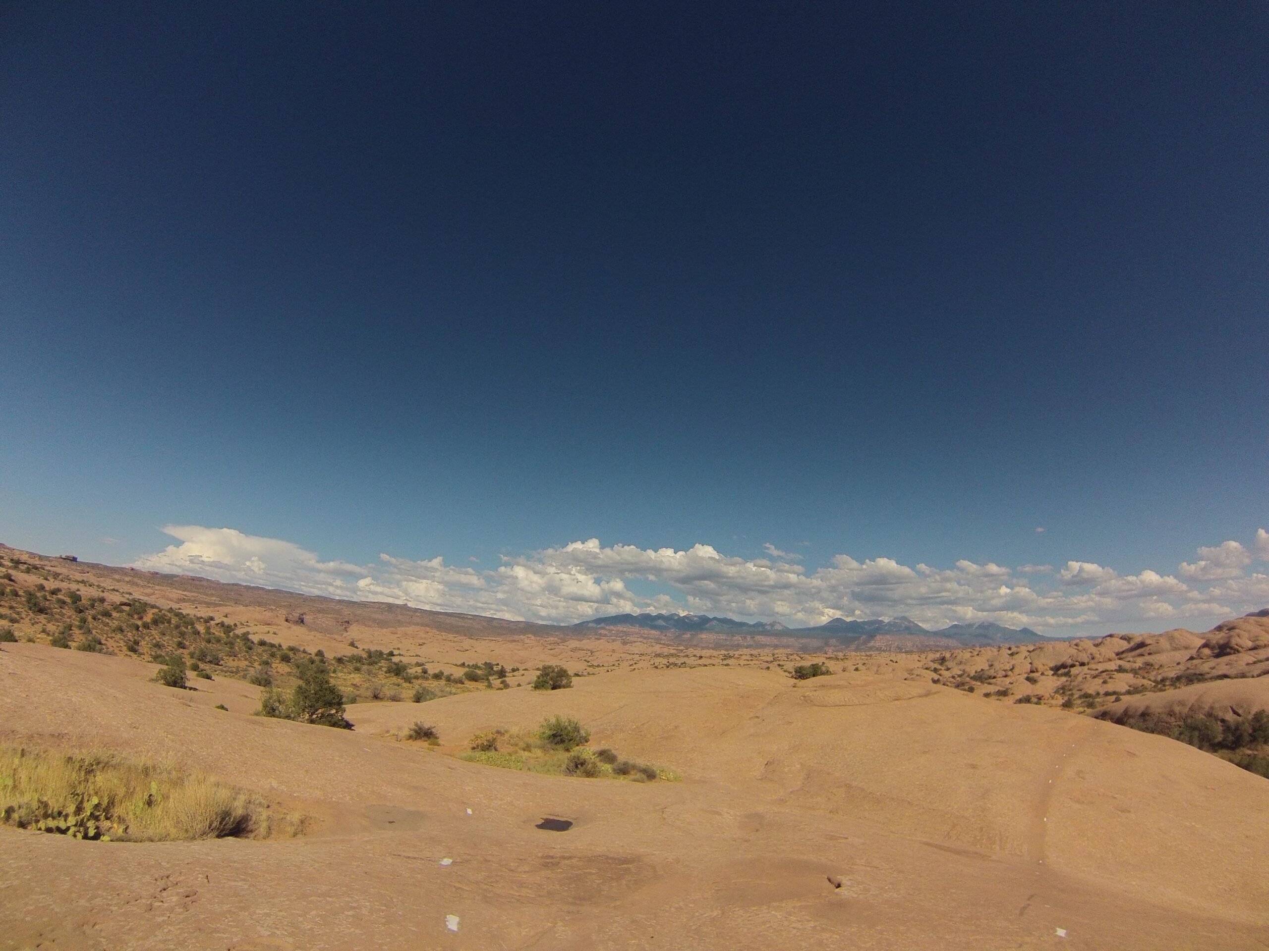 A vast desert landscape under a clear blue sky, featuring rolling sandy hills, scattered vegetation, and distant mountains. White clouds float in the sky, creating a contrast against the warm tones of the earth. Slickrock mountain bike trail.