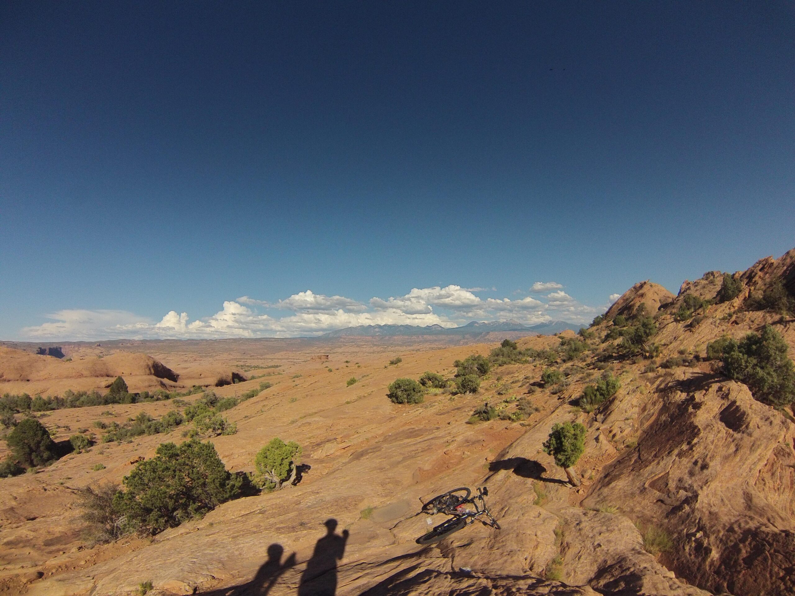 A panoramic view of a rugged, rocky landscape under a clear blue sky, featuring distant mountains and fluffy white clouds. In the foreground, the shadows of two people can be seen alongside a bicycle resting on the ground amidst sparse vegetation and rocky terrain. The scene captures the essence of outdoor adventure in a natural setting. Slickrock mountain bike trail.