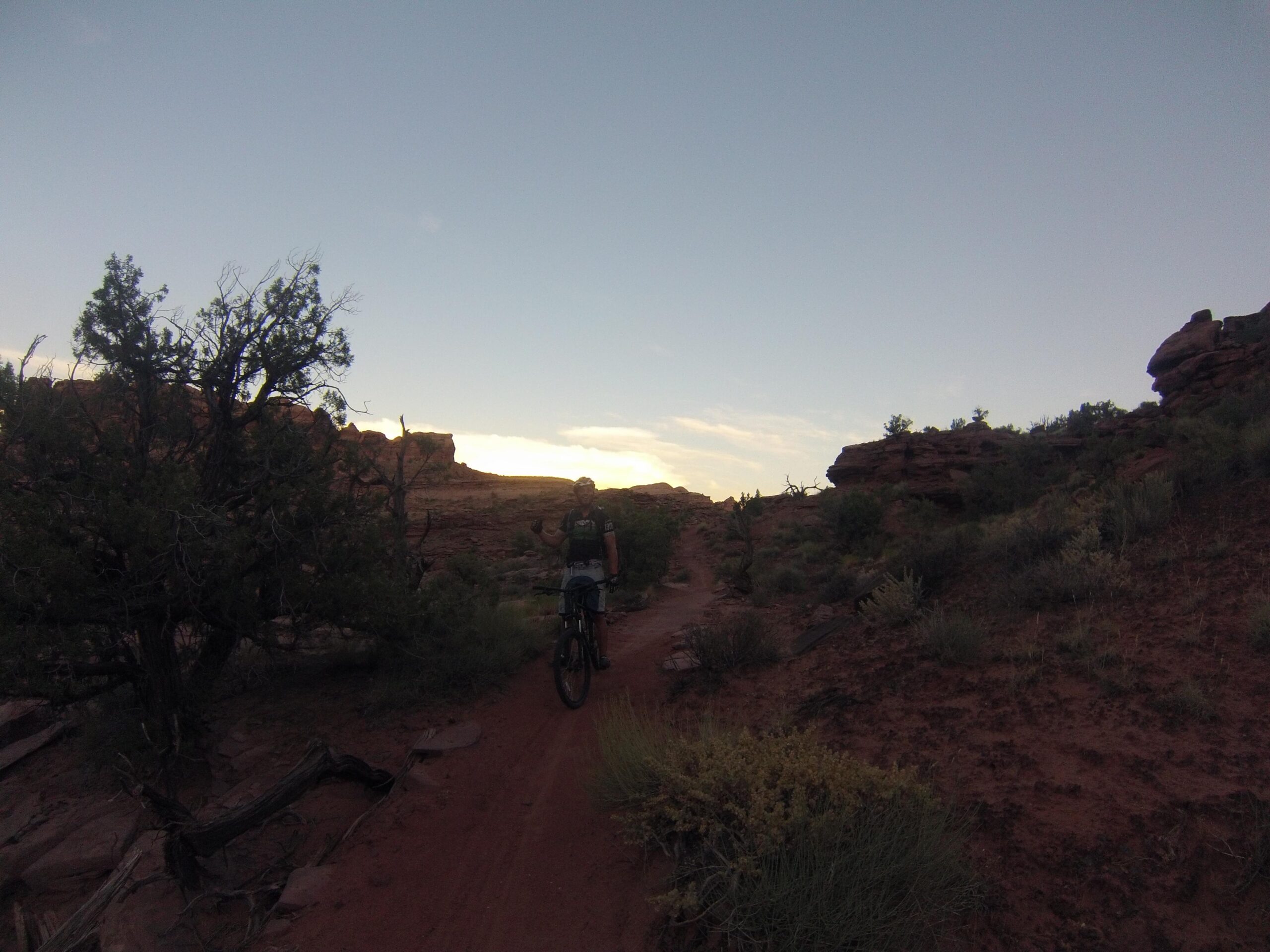 A person riding a mountain bike on a dirt trail surrounded by red rocks and sparse vegetation, with a clear sky and soft clouds in the background. Captain Ahab mountain bike trail.