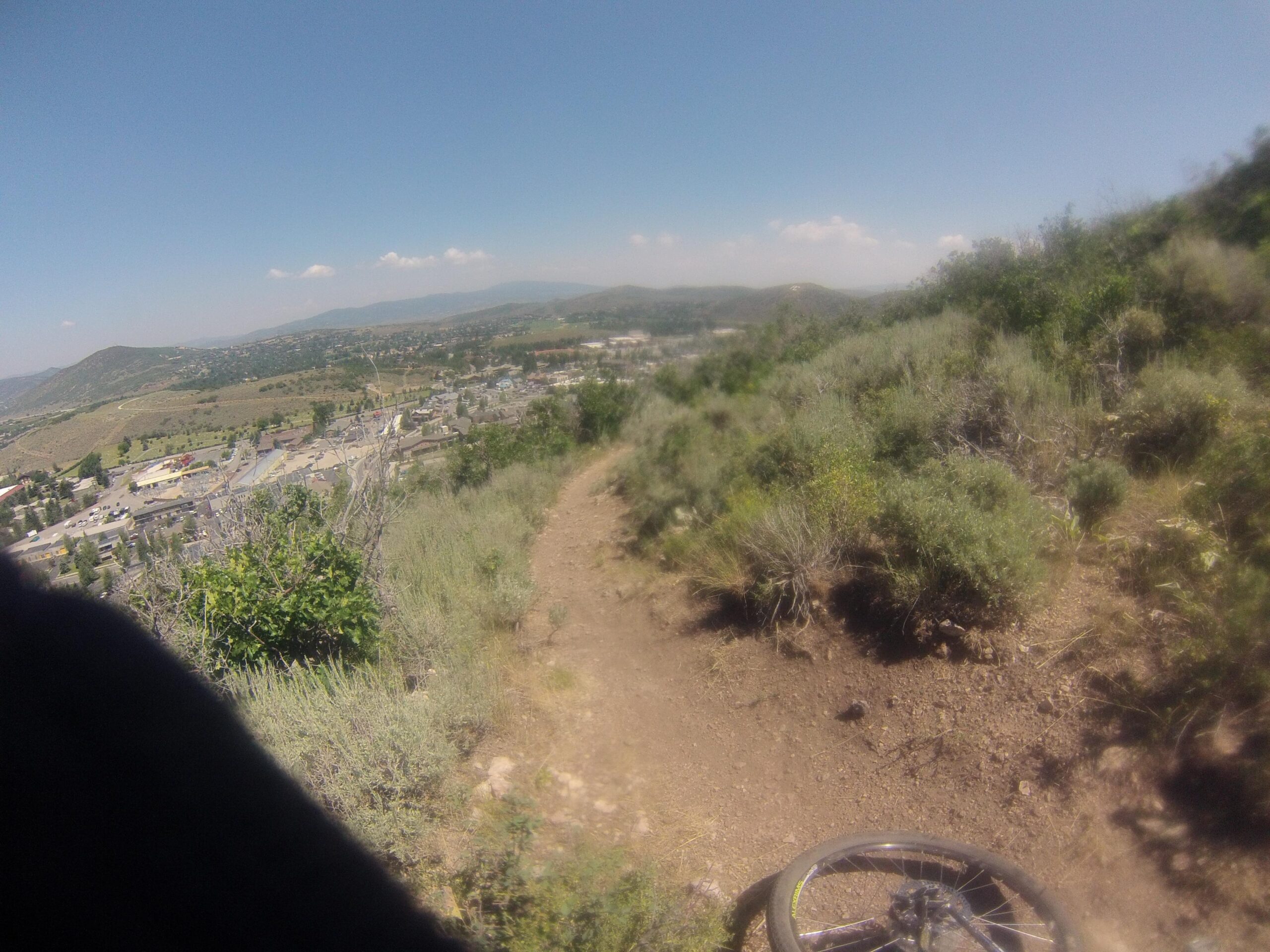 A view from a mountain biking trail overlooking a residential area, with hills in the background under a clear blue sky. The trail is surrounded by shrubs and small trees, with a bicycle wheel partially visible in the foreground. Masonic mountain bike trail.