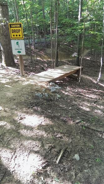 A wooden drop-off platform on a dirt trail in a wooded area, with a caution sign indicating "DROP." The sign includes symbols for mountain biking and recommends lowering speed. Surrounding trees and rocks are visible, suggesting a natural outdoor environment. Fountainhead Regional Park mountain bike trail.
