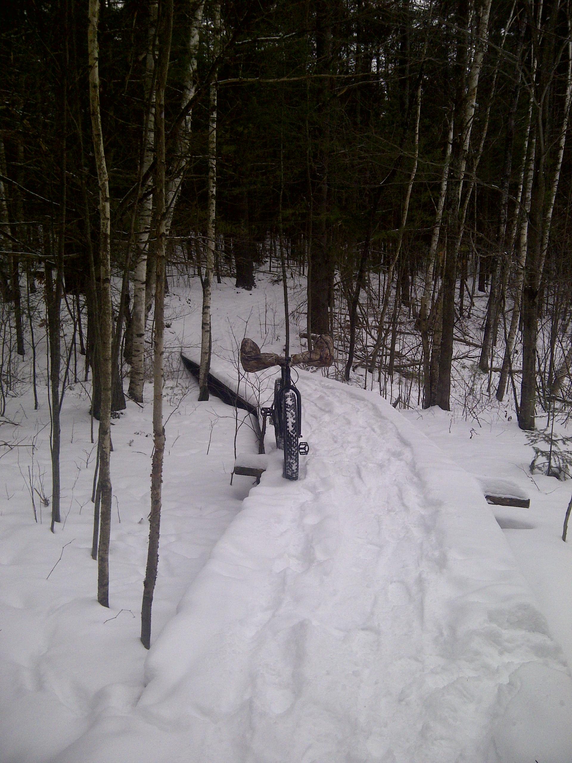 A snow-covered path winding through a forest of trees, with a bicycle parked on the side. The path is flanked by fresh snow, and wooden planks are visible, suggesting a bridge or raised walkway through the snowy landscape. The scene captures a quiet, wintery atmosphere. Ottawa River Corridor Trails mountain bike trail.