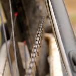 Close-up view of a bicycle chain running along the gears, with a slightly blurred background that includes parts of the bike frame. The image showcases the chain's links and the surrounding components, emphasizing the bike's mechanics.