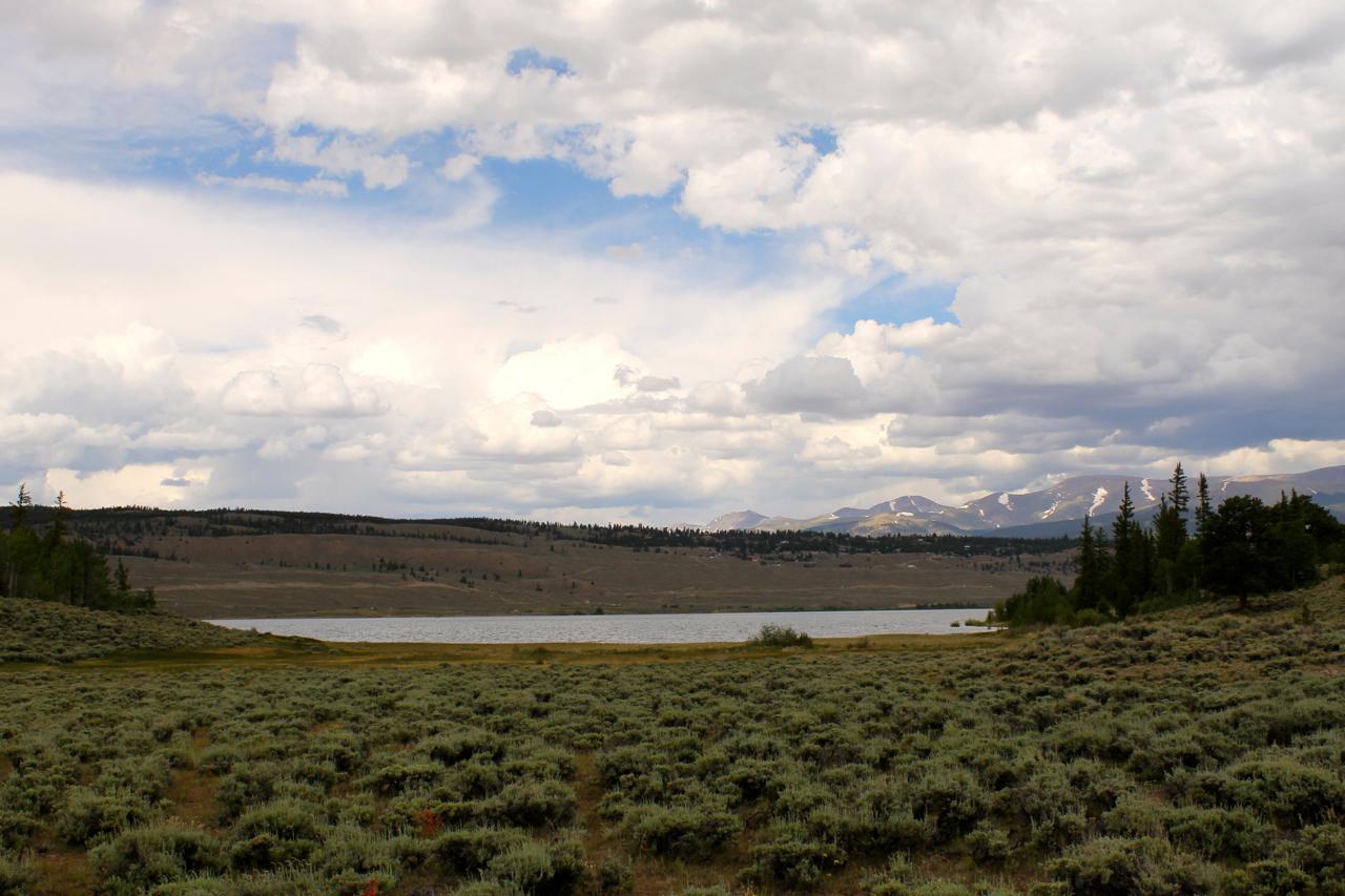 A serene landscape featuring a lake surrounded by rolling hills and sparse vegetation, under a mostly cloudy sky with patches of blue. In the background, snow-capped mountains can be seen, adding to the natural beauty of the scene. The foreground showcases a field of low shrubs, creating a peaceful and tranquil atmosphere. Twin Lakes Loop mountain bike trail.