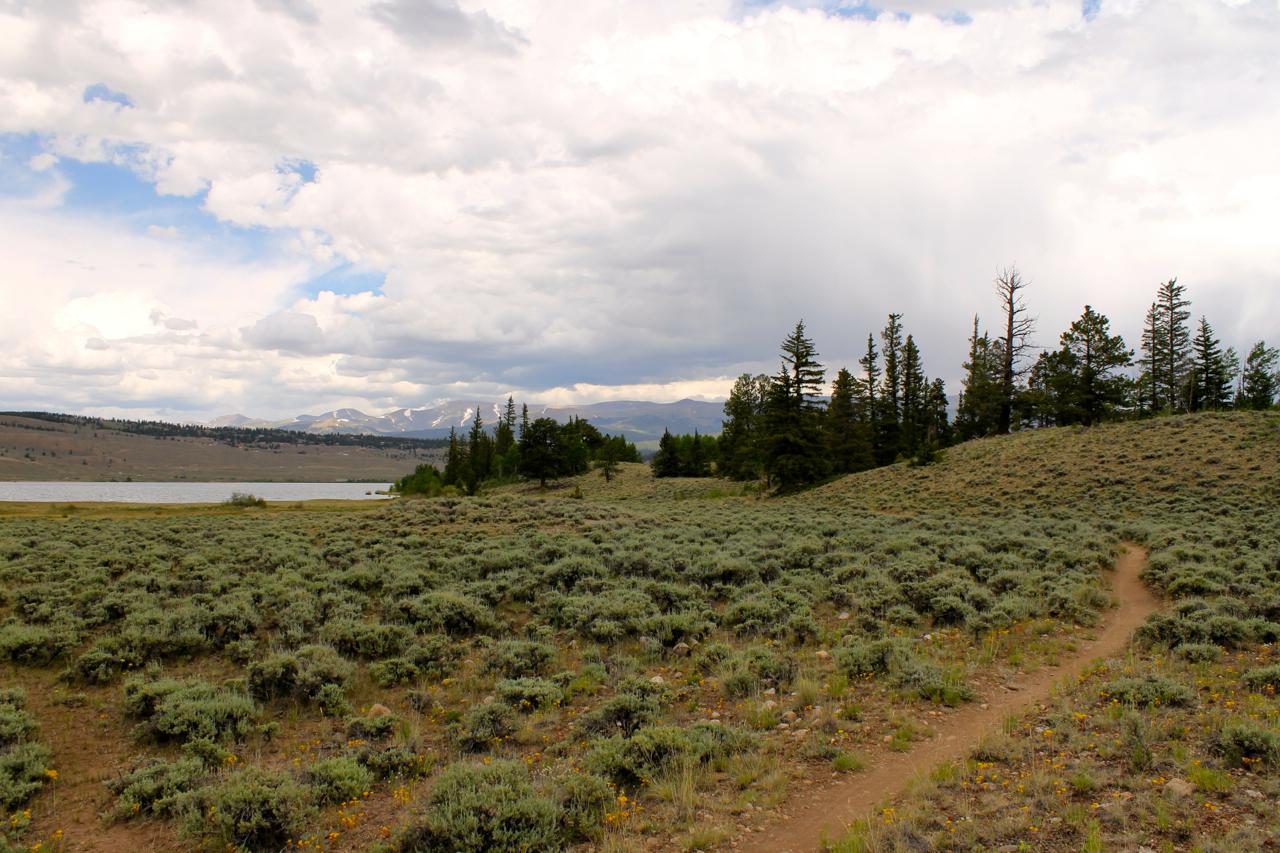 A scenic landscape featuring a grassy area with low shrubs, leading to a lake on the left. In the background, there are rolling hills and distant mountains under a sky filled with clouds. A dirt path winds through the foreground, inviting exploration. Twin Lakes Loop mountain bike trail.