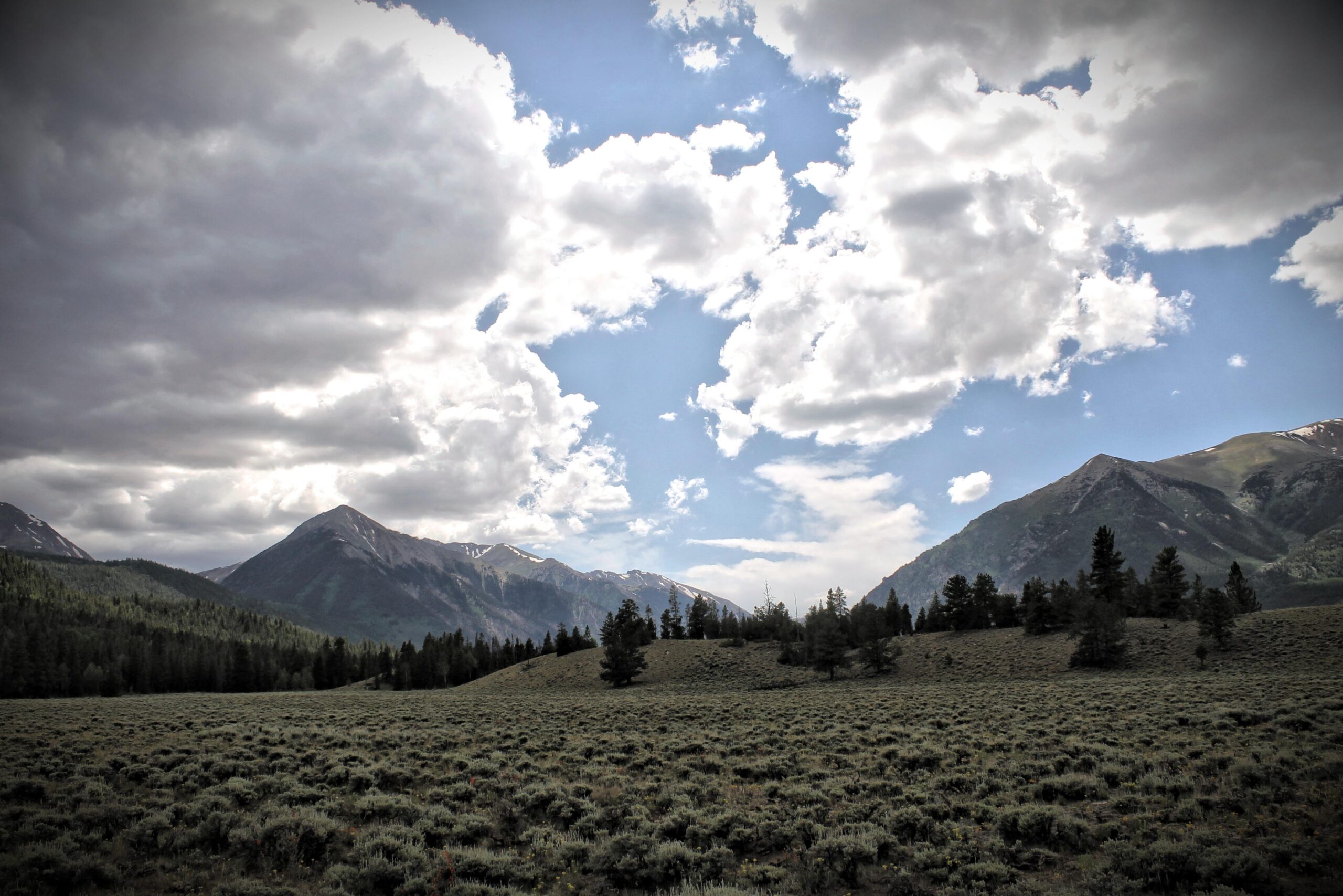 A scenic landscape featuring rolling hills covered in shrubs, with majestic mountains in the background beneath a partly cloudy sky. The lighting casts a serene atmosphere over the scene, highlighting the natural beauty of the outdoors. Twin Lakes Loop mountain bike trail.