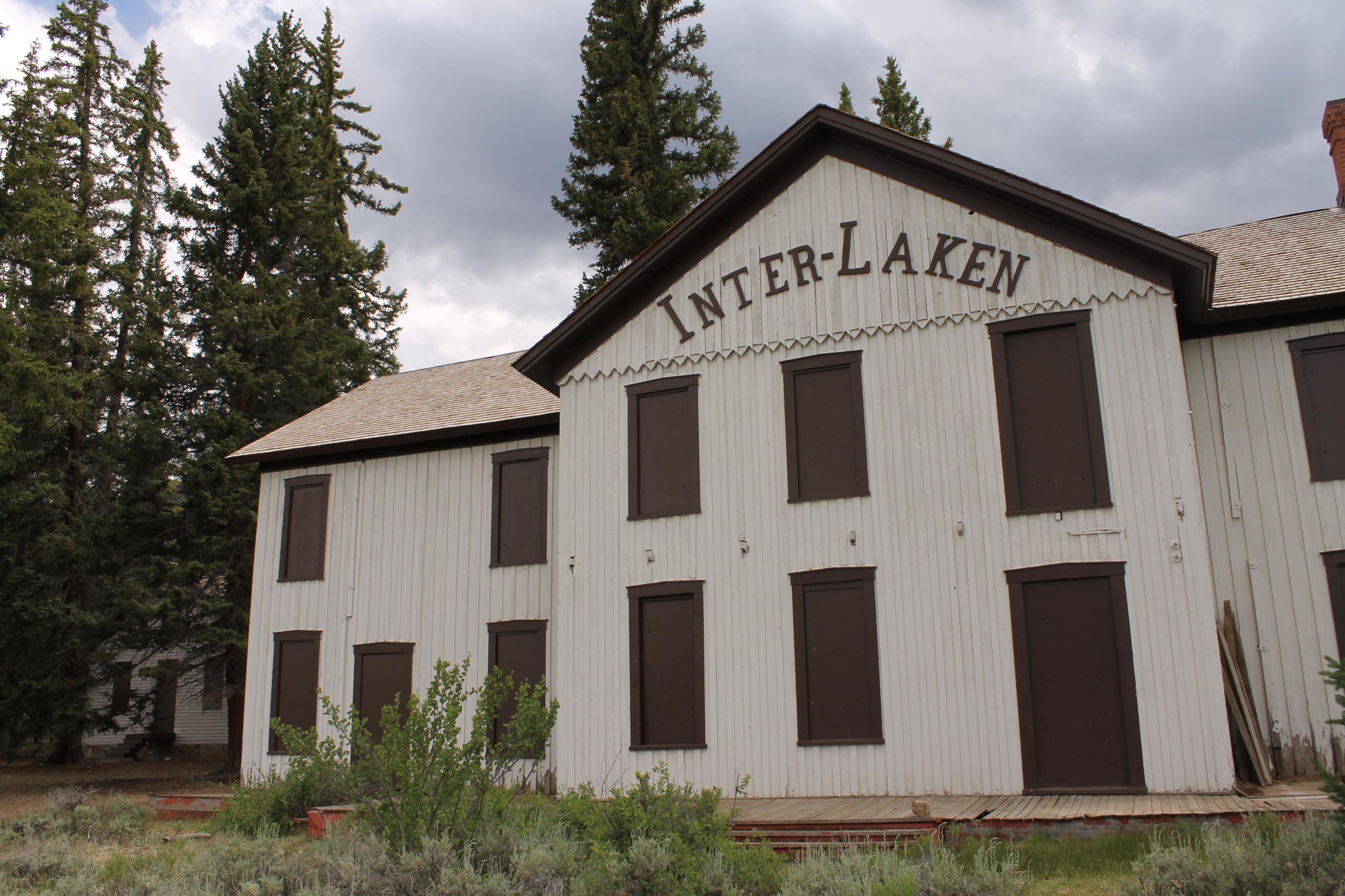An old wooden building named "Inter-Laken" with boarded-up windows, surrounded by tall pine trees and a cloudy sky. The structure features white siding with brown trim and a sloping facade, giving it a rustic and abandoned appearance. Wild greenery is visible in front of the building. Colorado Trail: Clear Creek Thd to Lake View CG / Hwy 82 mountain bike trail.
