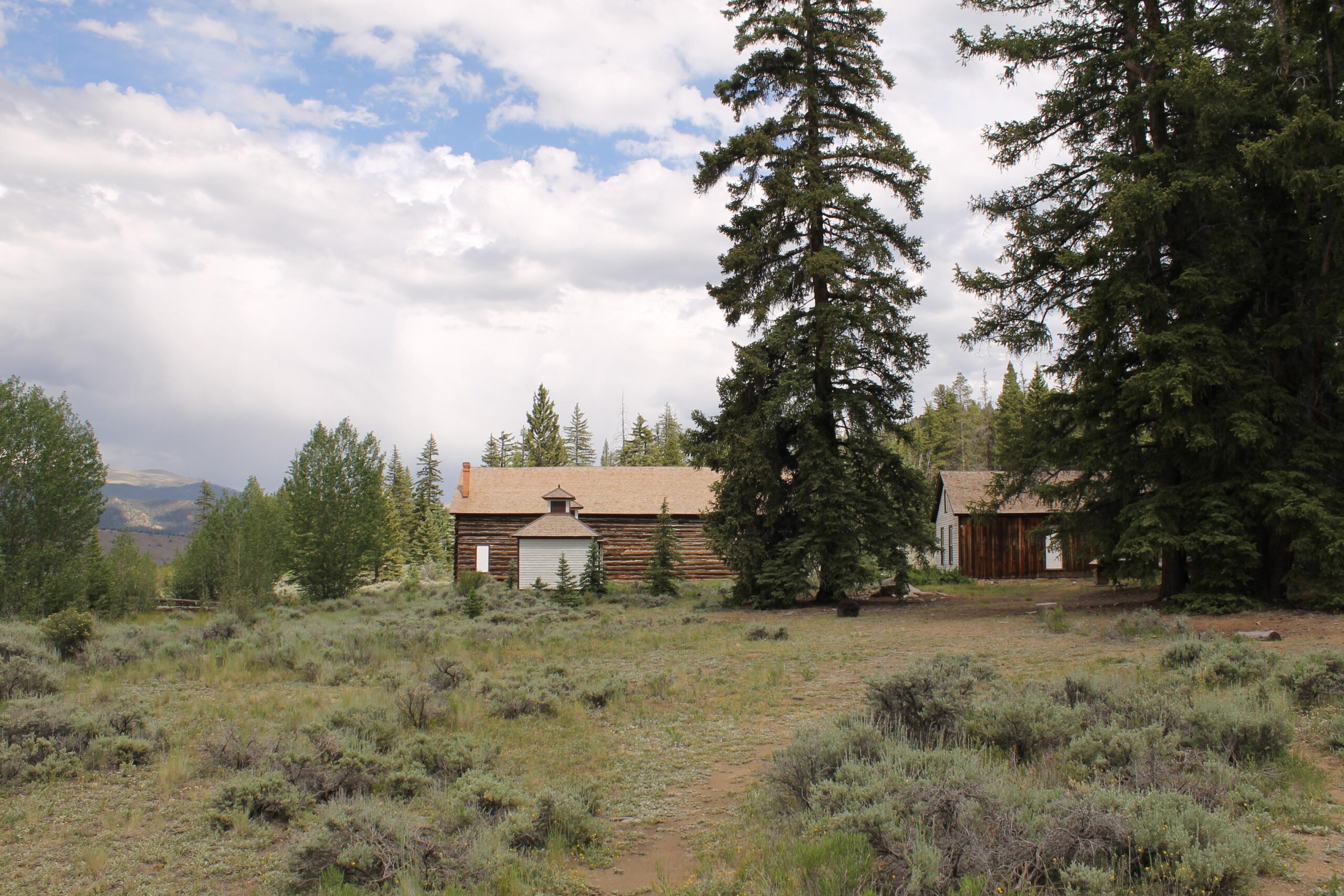 A rustic cabin surrounded by tall pine trees and sagebrush, set against a cloudy sky. The cabin features a log exterior with a sloped roof and a small structure attached to its front. In the background, rolling mountains are visible through the trees. The landscape is lush and green, indicating a natural setting. Twin Lakes Loop mountain bike trail.