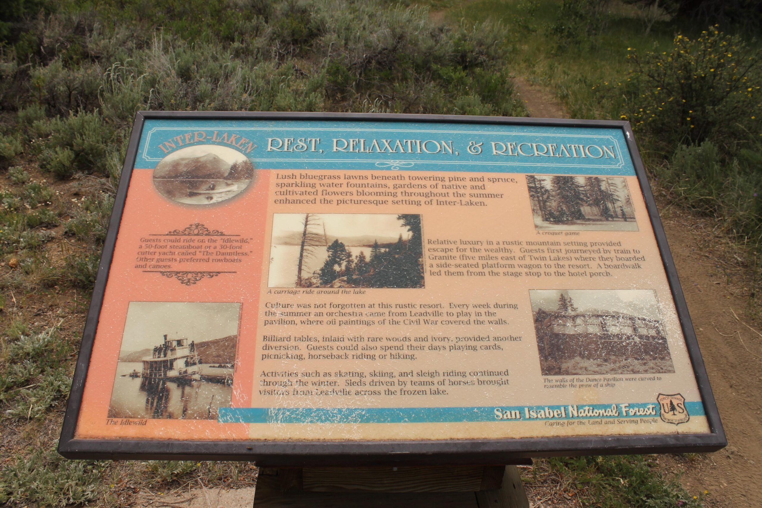 Sign describing the historical resort of Inter-Laken, featuring information about its amenities, leisure activities, and cultural events. The sign includes illustrations of the resort's attractions, such as a steamboat and a croquet game. Surrounded by natural foliage, it provides a glimpse into the past, highlighting the picturesque setting and recreational options available to guests. Twin Lakes Loop mountain bike trail.