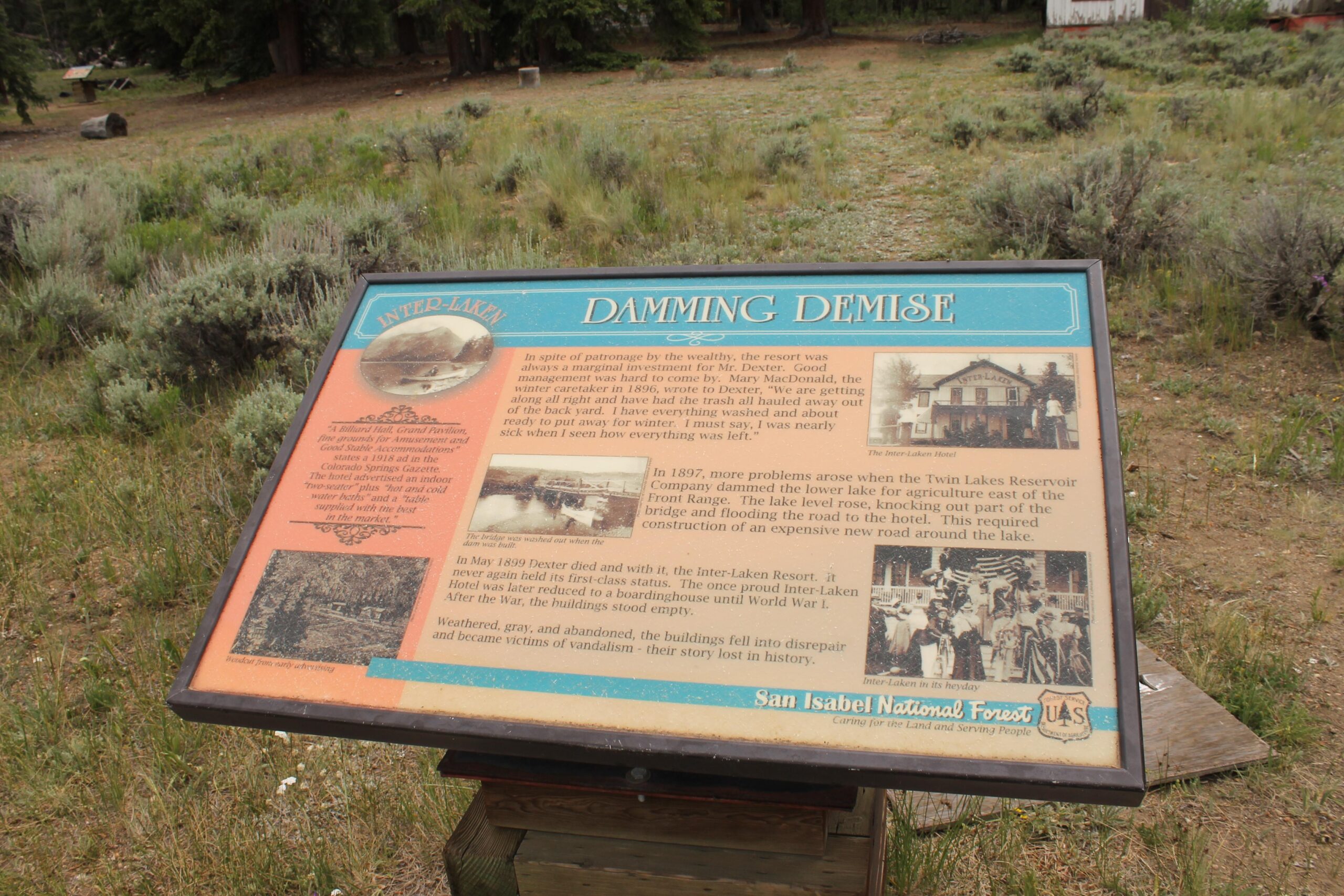 A wooden informational sign titled "Damming Demise" in the San Isabel National Forest, detailing the history of the Inter-Laken Resort. The sign includes images and text describing the resort's challenges, including damming issues affecting the surrounding lakes, and mentions its decline and eventual abandonment. The background features a mostly grassy area with sparse shrubs and trees. Twin Lakes Loop mountain bike trail.