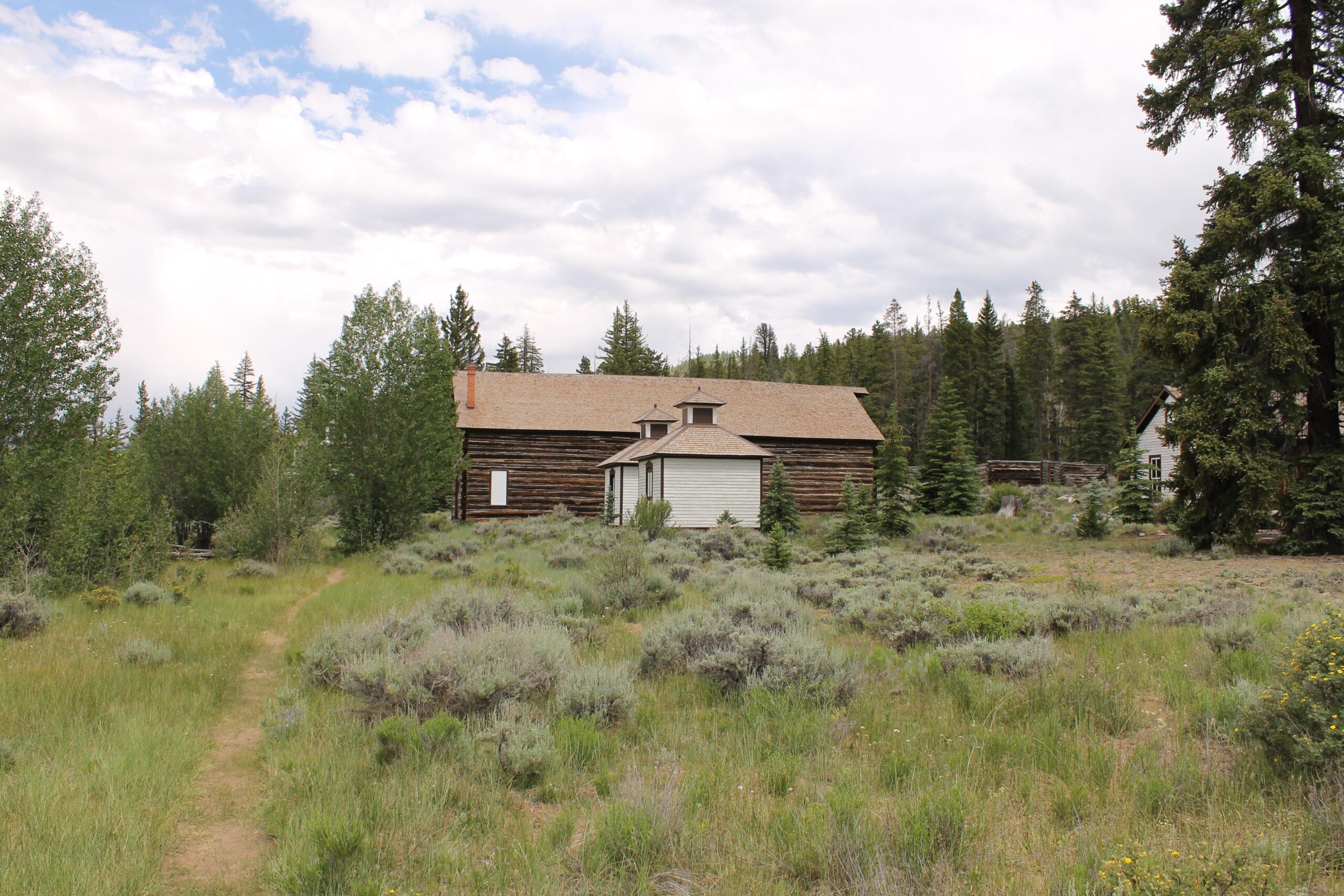 A rustic log cabin surrounded by lush greenery and small shrubs, with a dirt path leading through the tall grass. The cabin features a large front porch and a peaked roof, set against a backdrop of evergreen trees and a partly cloudy sky. Twin Lakes Loop mountain bike trail.