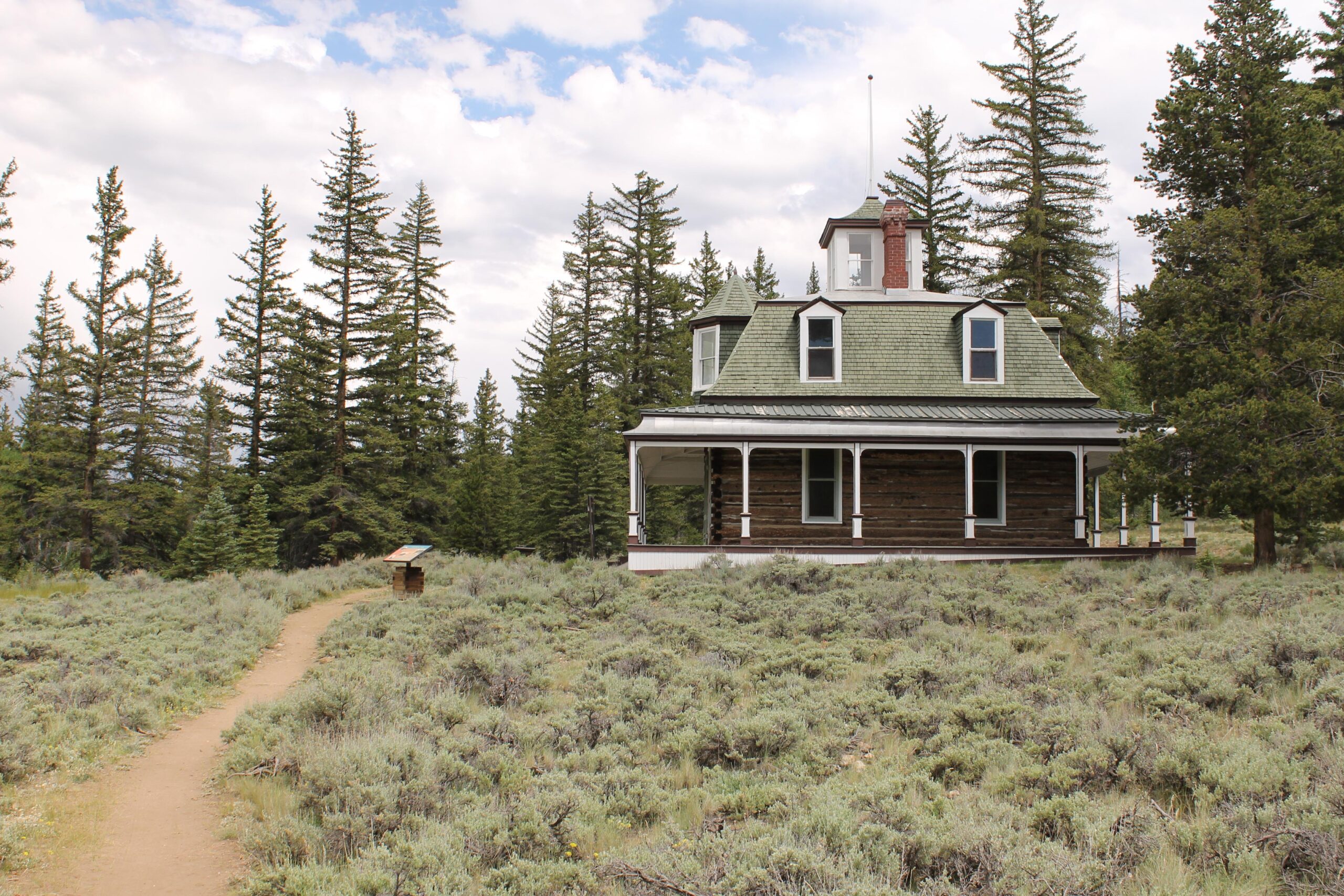A rustic wooden house with a green roof situated in a grassy area surrounded by tall pine trees under a partly cloudy sky. A dirt path leads to the house. Twin Lakes Loop mountain bike trail.