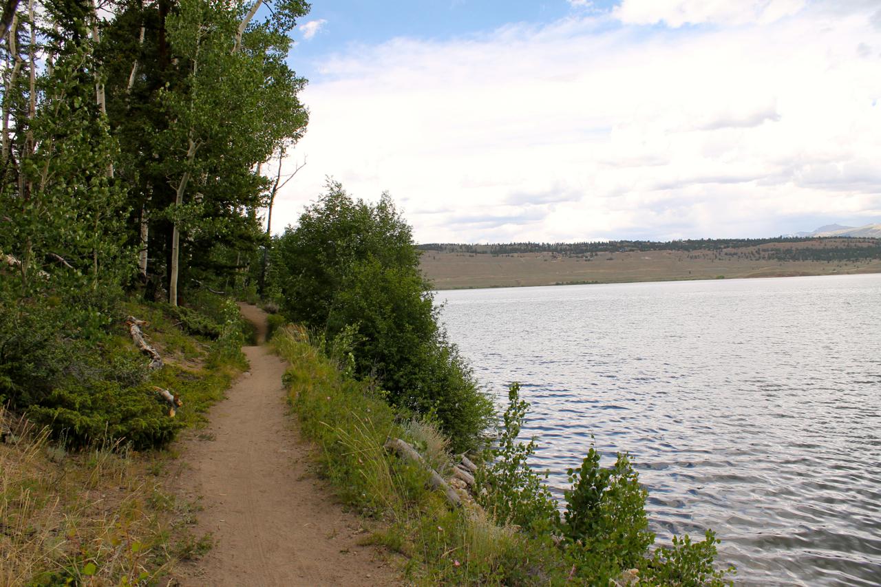 A scenic dirt pathway lined with greenery runs alongside a calm lake, with gentle hills visible in the background beneath a partly cloudy sky. Twin Lakes Loop mountain bike trail.