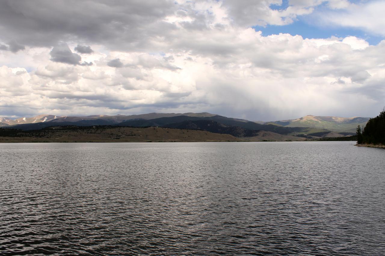 A tranquil lake surrounded by rolling hills and mountains under a cloudy sky. The water reflects the clouds, creating a serene atmosphere. Twin Lakes Loop mountain bike trail.