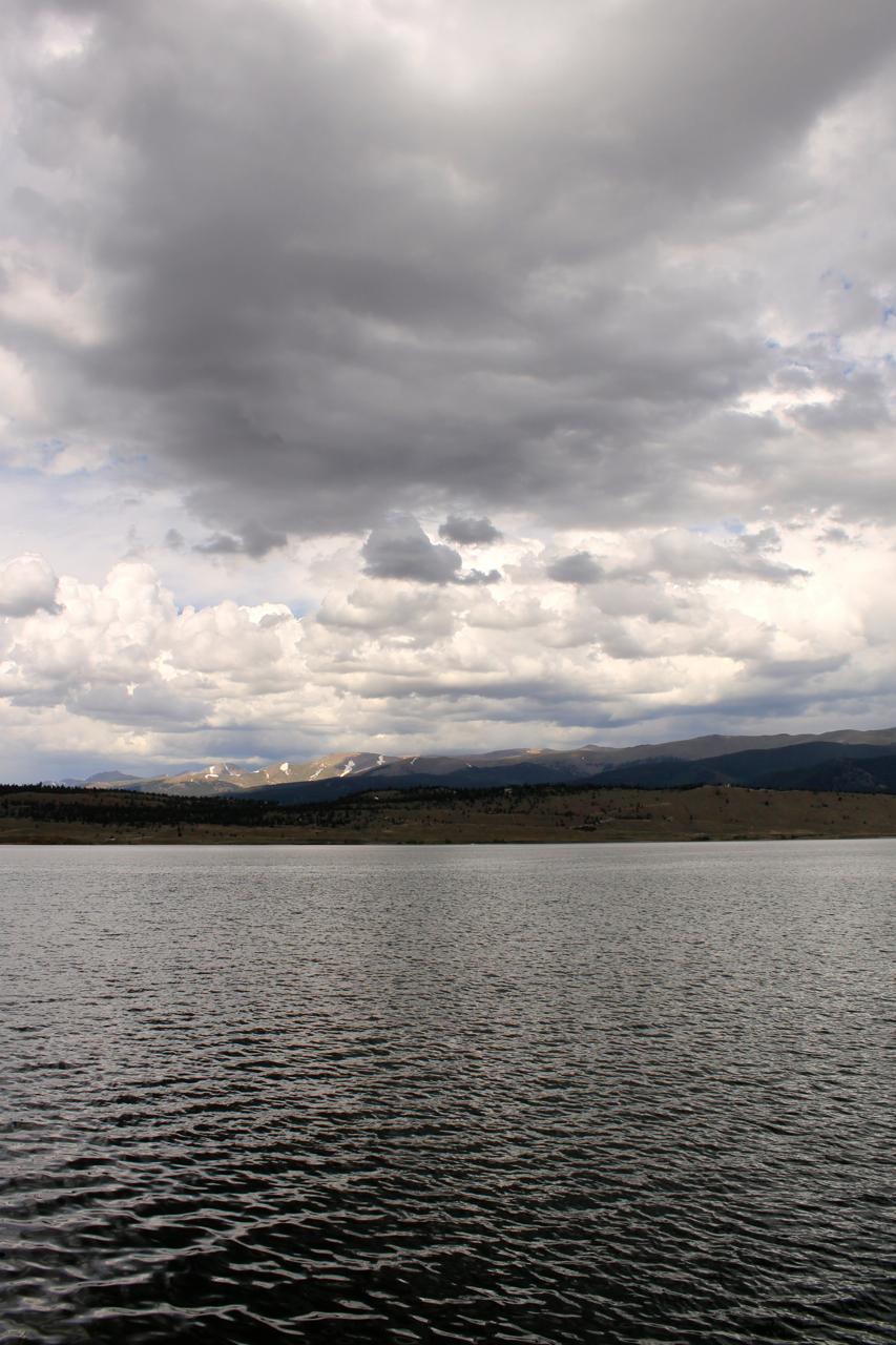 A serene view of a lake reflecting the overcast sky, with dramatic clouds above and mountains in the distance. The water surface is slightly rippled, suggesting a gentle breeze. Colorado Trail: Clear Creek Thd to Lake View CG / Hwy 82 mountain bike trail.