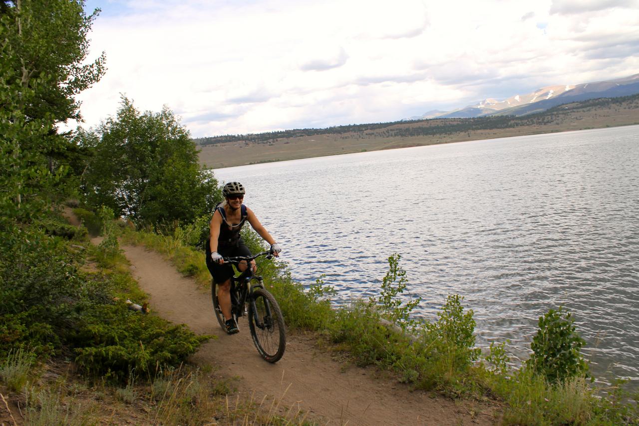 A person riding a mountain bike along a dirt path next to a lake, surrounded by greenery and trees, with mountains visible in the background under a partly cloudy sky. Colorado Trail: Clear Creek Thd to Lake View CG / Hwy 82 mountain bike trail.