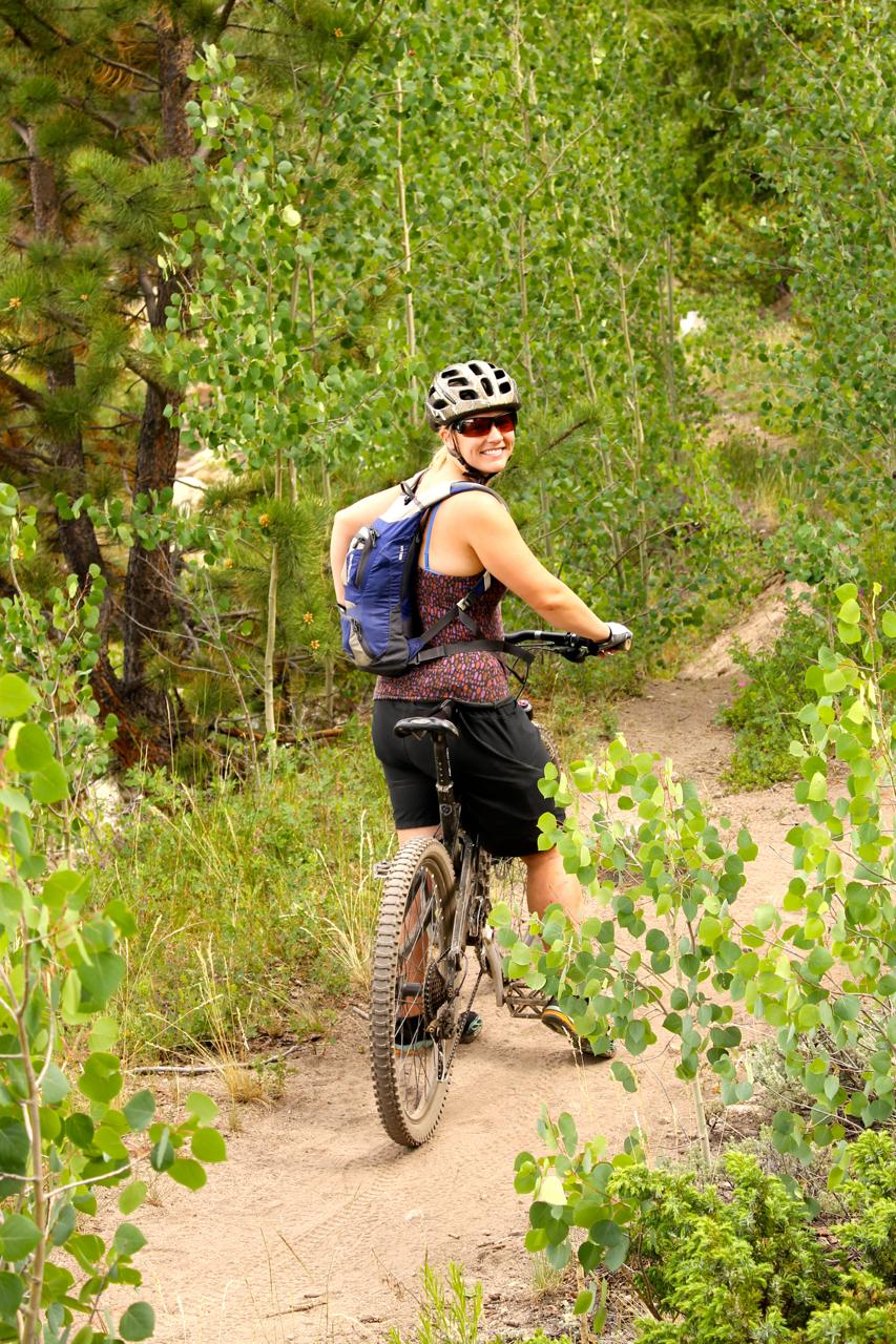 A woman on a mountain bike smiles over her shoulder while riding along a narrow dirt trail surrounded by green foliage and aspen trees. She wears a helmet and sunglasses and has a blue backpack. The scene captures a sunny day and a sense of outdoor adventure. Colorado Trail: Clear Creek Thd to Lake View CG / Hwy 82 mountain bike trail.