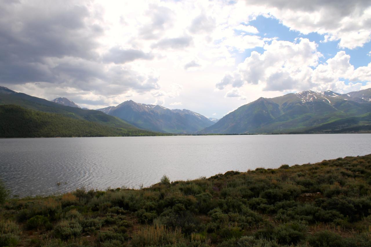 A scenic view of a tranquil lake surrounded by lush green hills and majestic mountains under a partly cloudy sky. The water reflects the surrounding landscape, creating a serene atmosphere. Twin Lakes Loop mountain bike trail.