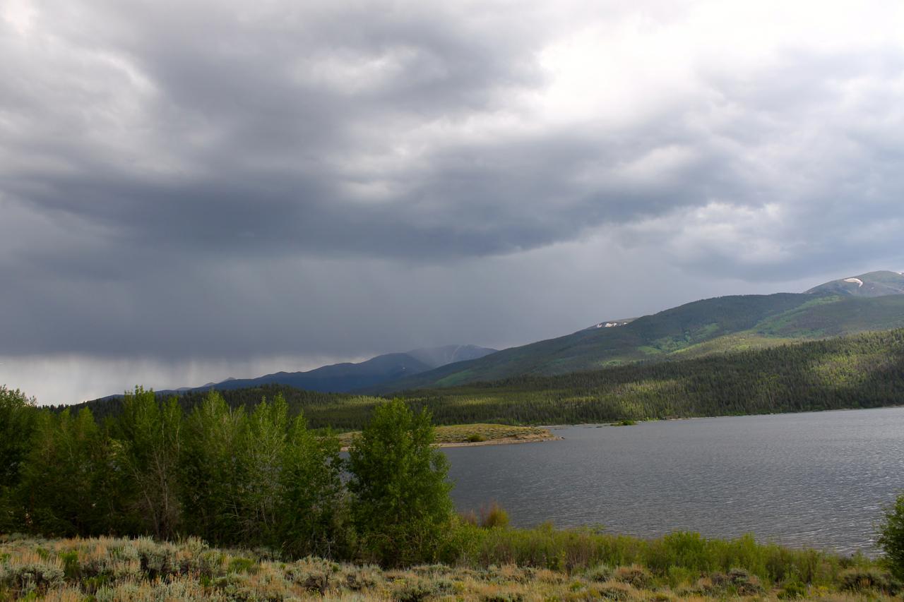 A scenic landscape featuring a calm lake surrounded by lush green trees and rolling mountains under a dramatic sky filled with dark clouds. Light rain can be seen falling in the distance, suggesting an impending storm. Colorado Trail: Clear Creek Thd to Lake View CG / Hwy 82 mountain bike trail.