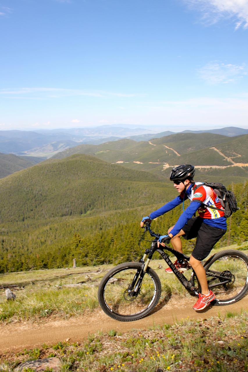A person riding a mountain bike on a dirt trail, surrounded by lush green hills and trees, with a clear blue sky above and distant mountains in the background. Monarch Crest Trail mountain bike trail.