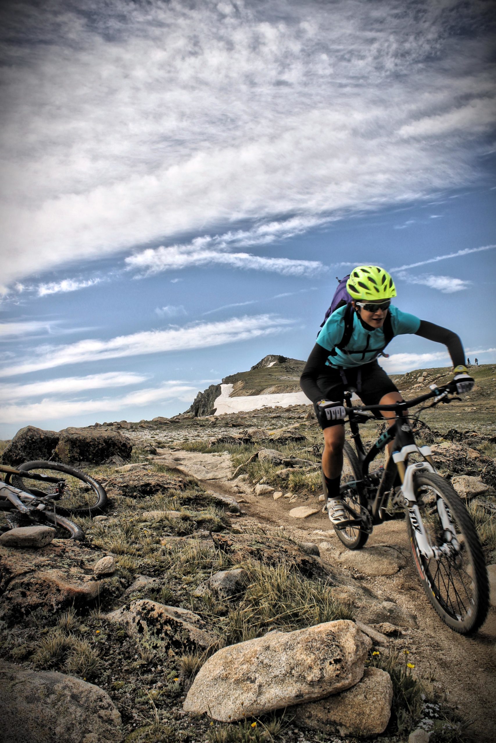 A mountain biker navigating a rocky trail in a mountainous area, with a clear blue sky and wispy clouds in the background. The biker is wearing a bright yellow helmet and a light blue shirt, focused on the path ahead. An abandoned bicycle is visible in the foreground, surrounded by rocky terrain and sparse vegetation. Monarch Crest Trail mountain bike trail.