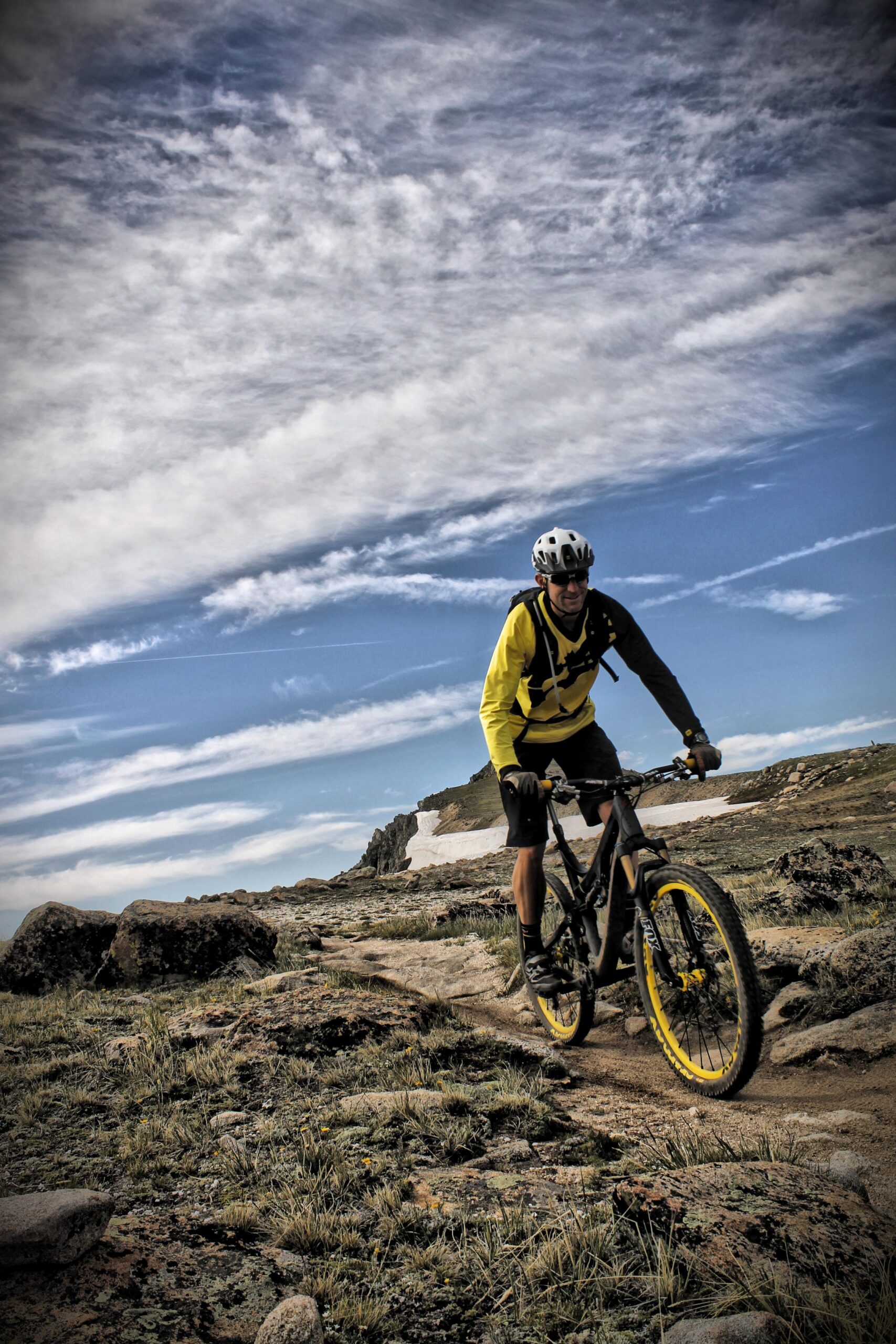 A mountain biker wearing a yellow and black outfit rides along a rocky trail under a blue sky with scattered clouds. The landscape features rugged terrain and patches of snow in the background. Monarch Crest Trail mountain bike trail.