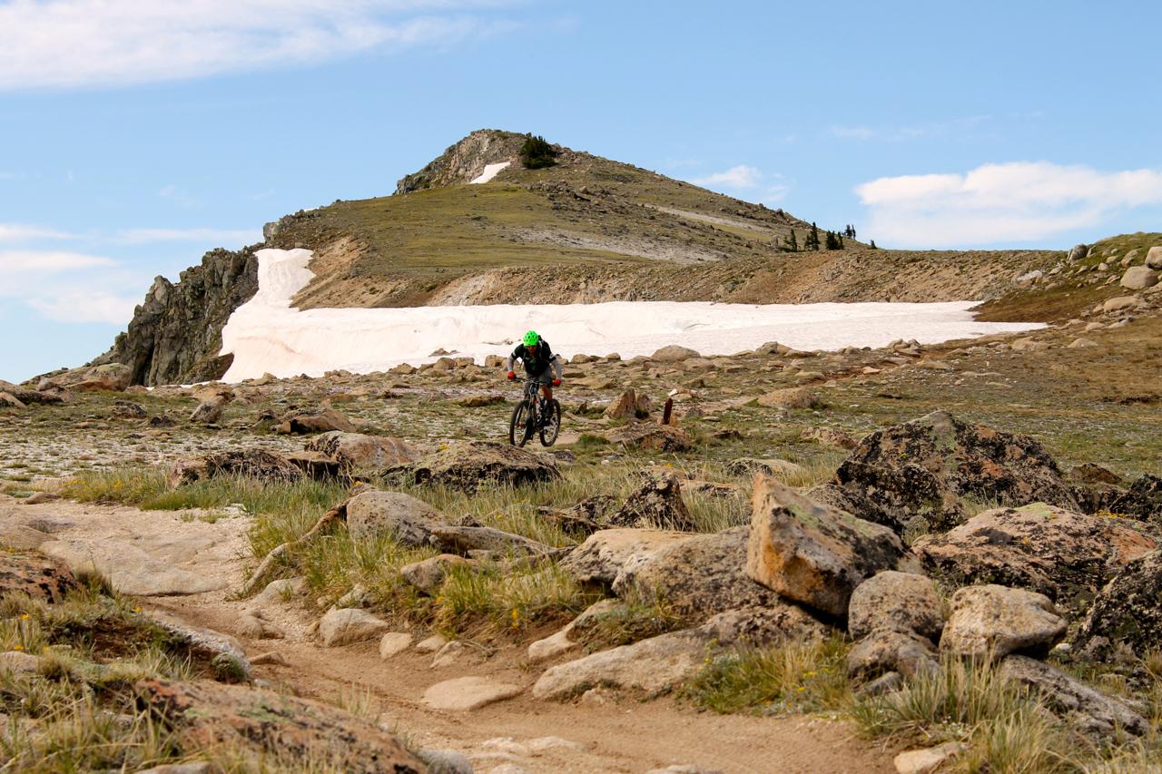 A mountain biker navigates a rocky terrain with a snowy slope in the background under a blue sky with scattered clouds. Monarch Crest Trail mountain bike trail.