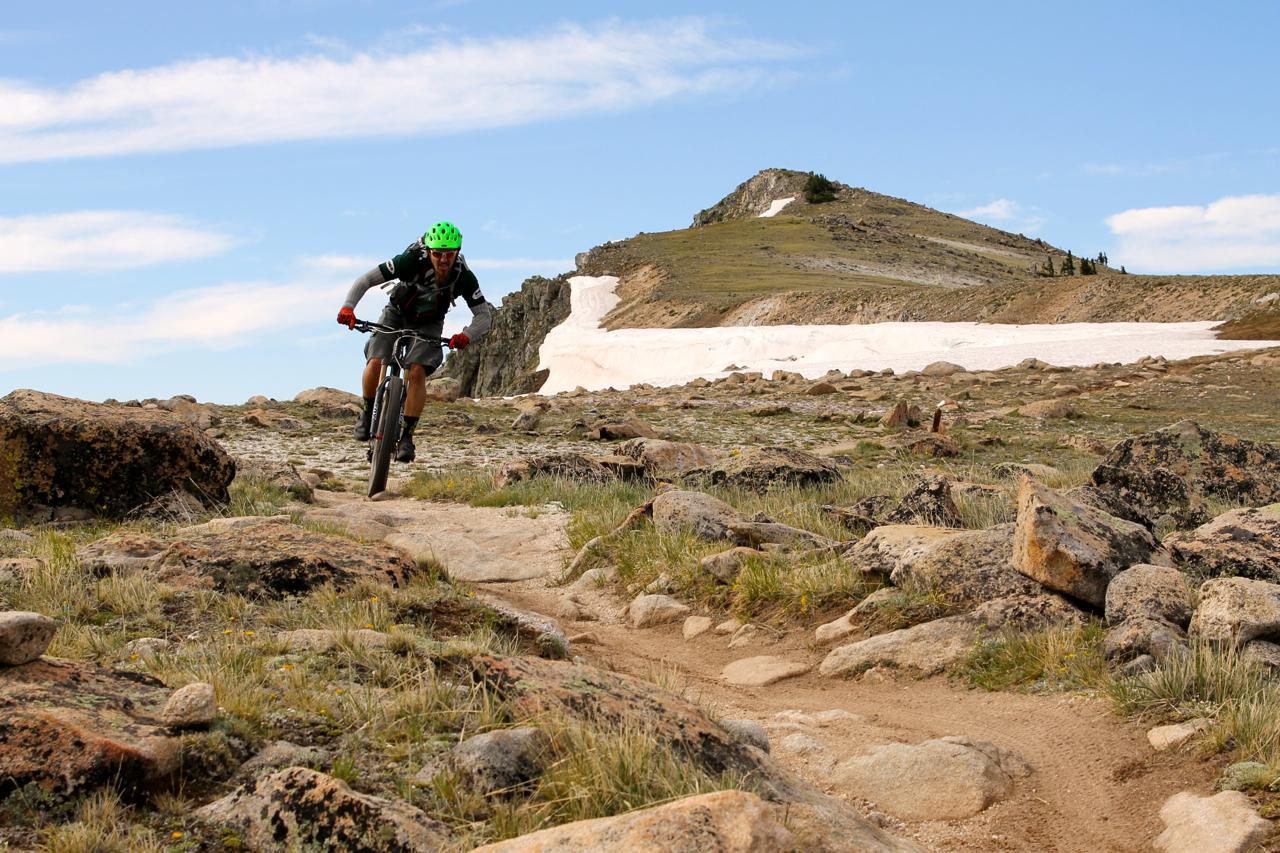 A mountain biker riding along a rocky trail with snow-covered ground in the background, under a blue sky with scattered clouds. Monarch Crest Trail mountain bike trail.