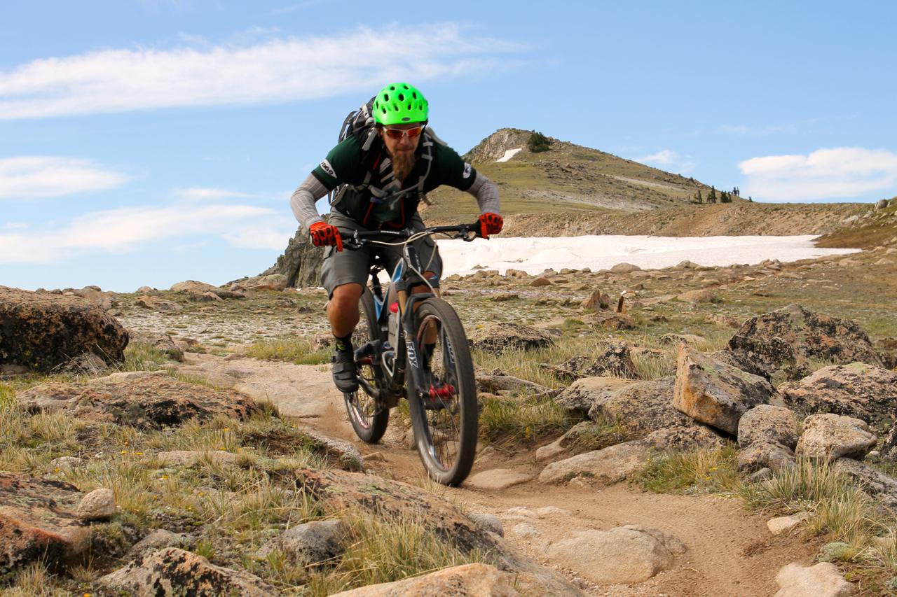 A mountain biker in a green helmet navigates a rocky trail surrounded by grass and boulders, with snow-capped hills in the background under a blue sky with clouds. Monarch Crest Trail mountain bike trail.