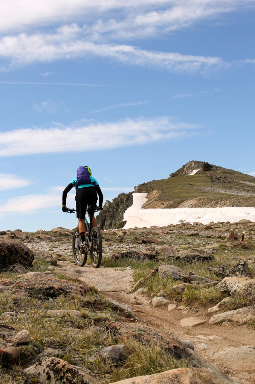 A cyclist rides along a rocky mountain trail under a blue sky with wispy clouds. Snow can be seen on the hillside in the background, and the landscape features sparse grass and large stones. The cyclist is dressed in a blue and green jacket with a purple backpack, pedaling away from the camera. Monarch Crest Trail mountain bike trail.