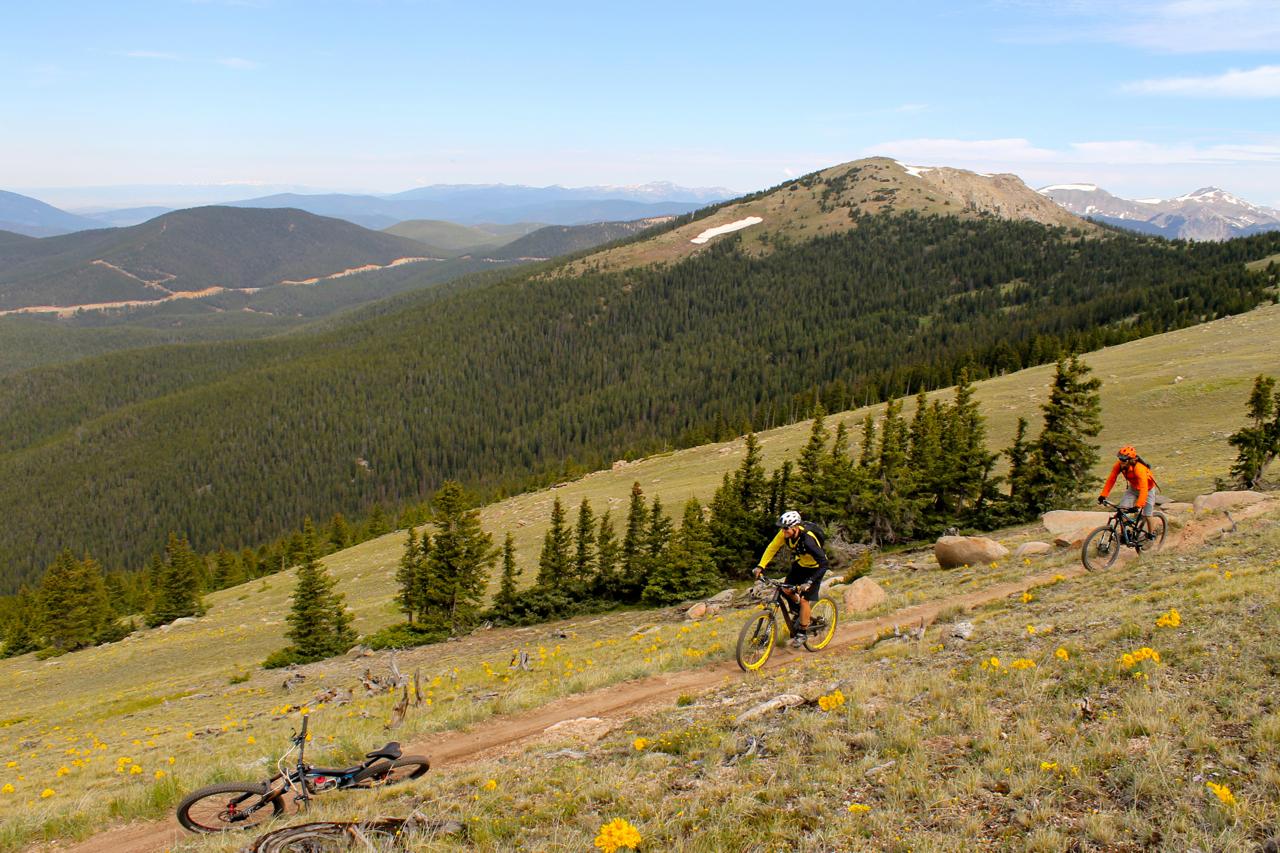 Two mountain bikers navigating a dirt trail on a hillside surrounded by dense greenery and distant mountains. The landscape features patches of wildflowers and rocky outcrops under a clear blue sky. One biker is wearing a yellow shirt, while the other is in an orange shirt. Monarch Crest Trail mountain bike trail.