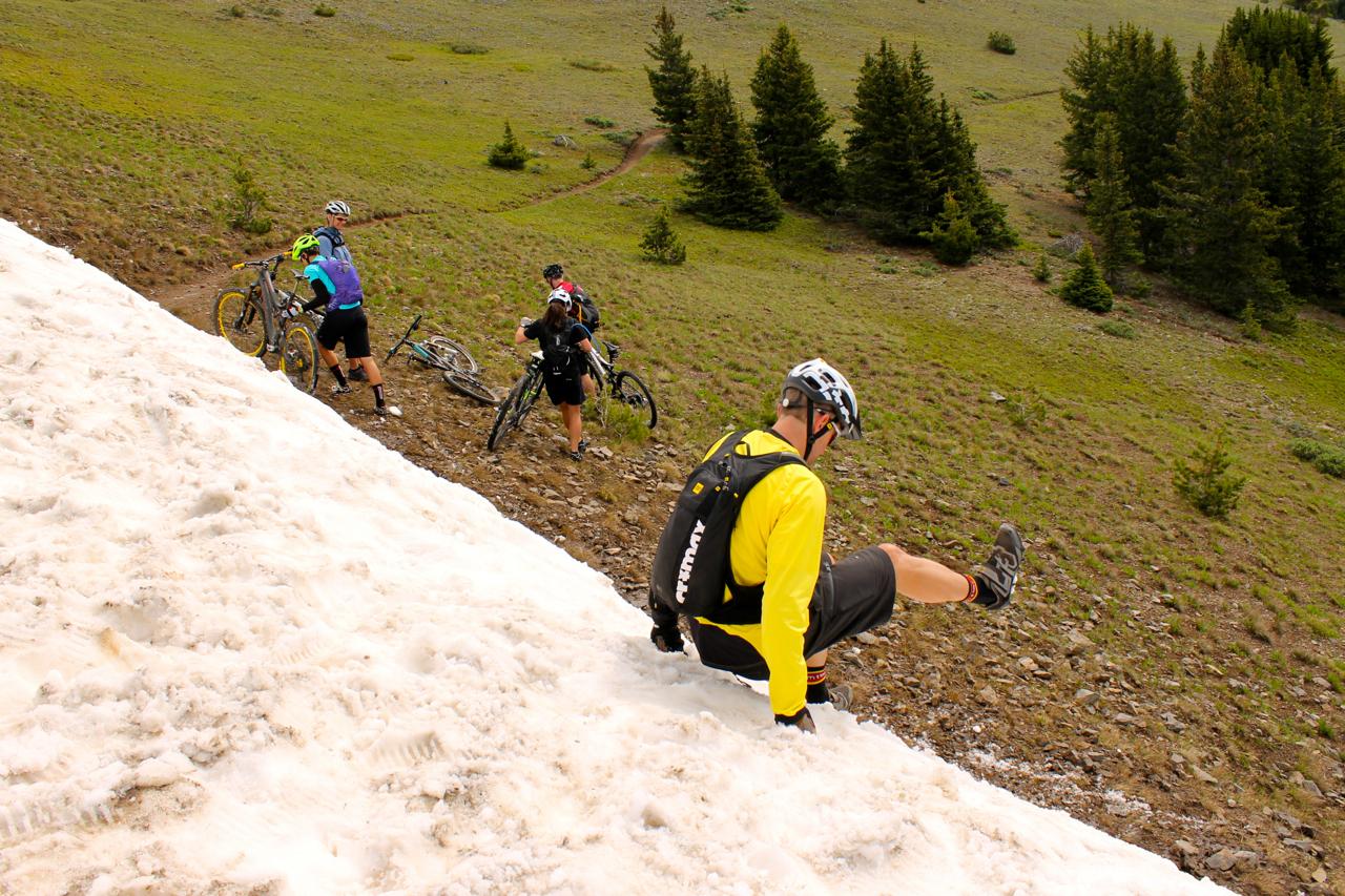 A group of mountain bikers navigating a snowy slope in a mountain area. One rider is sliding down the snow while others are pushing their bikes uphill. The landscape features green grass and trees in the background, indicating a mix of winter and spring conditions. Monarch Crest Trail mountain bike trail.