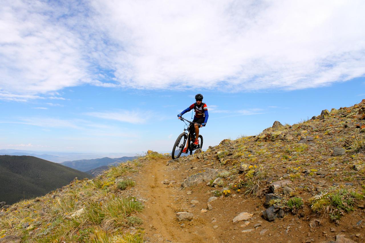 A mountain biker navigating a rocky trail on a hillside, surrounded by green grass and wildflowers under a blue sky with scattered clouds. The landscape features rolling mountains in the background. Monarch Crest Trail mountain bike trail.