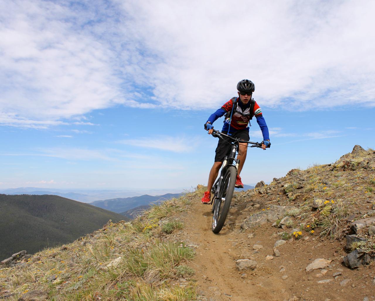 A person riding a mountain bike on a narrow dirt trail along a hillside, surrounded by rocky terrain and sparse vegetation. The sky is partly cloudy, with blue patches visible. The rider is wearing a helmet and a colorful cycling outfit. Monarch Crest Trail mountain bike trail.