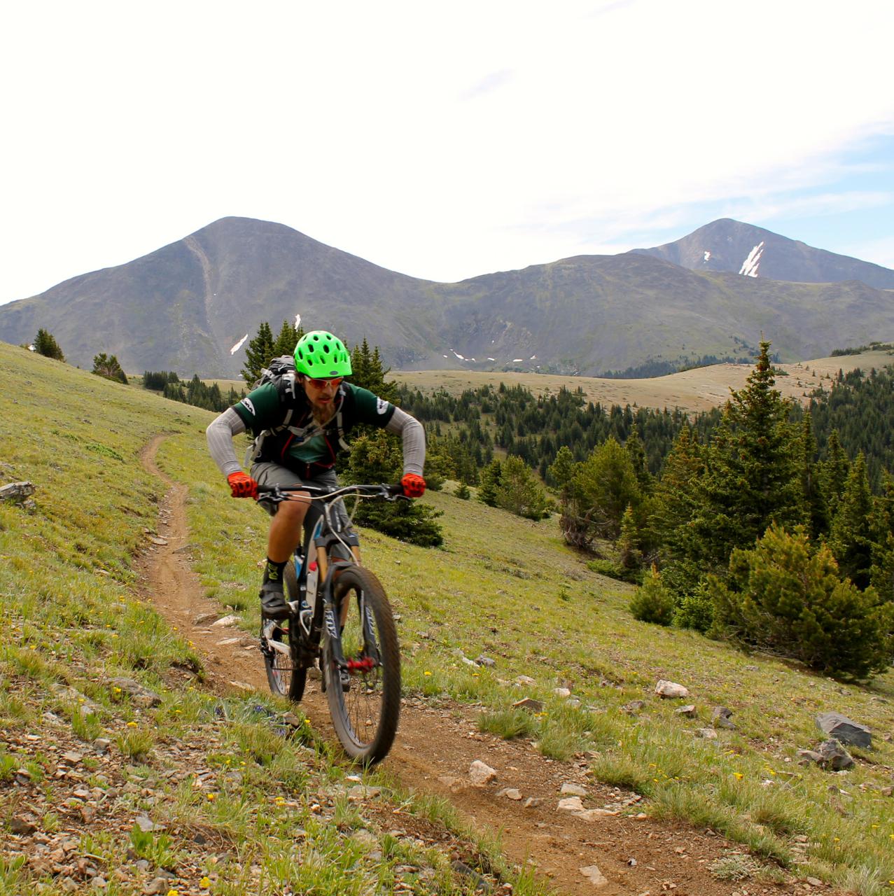 A person riding a mountain bike along a dirt trail on a grassy hillside, with mountains in the background and trees lining the path. The cyclist is wearing a bright green helmet and orange gloves, and is focused on navigating the terrain. Monarch Crest Trail mountain bike trail.